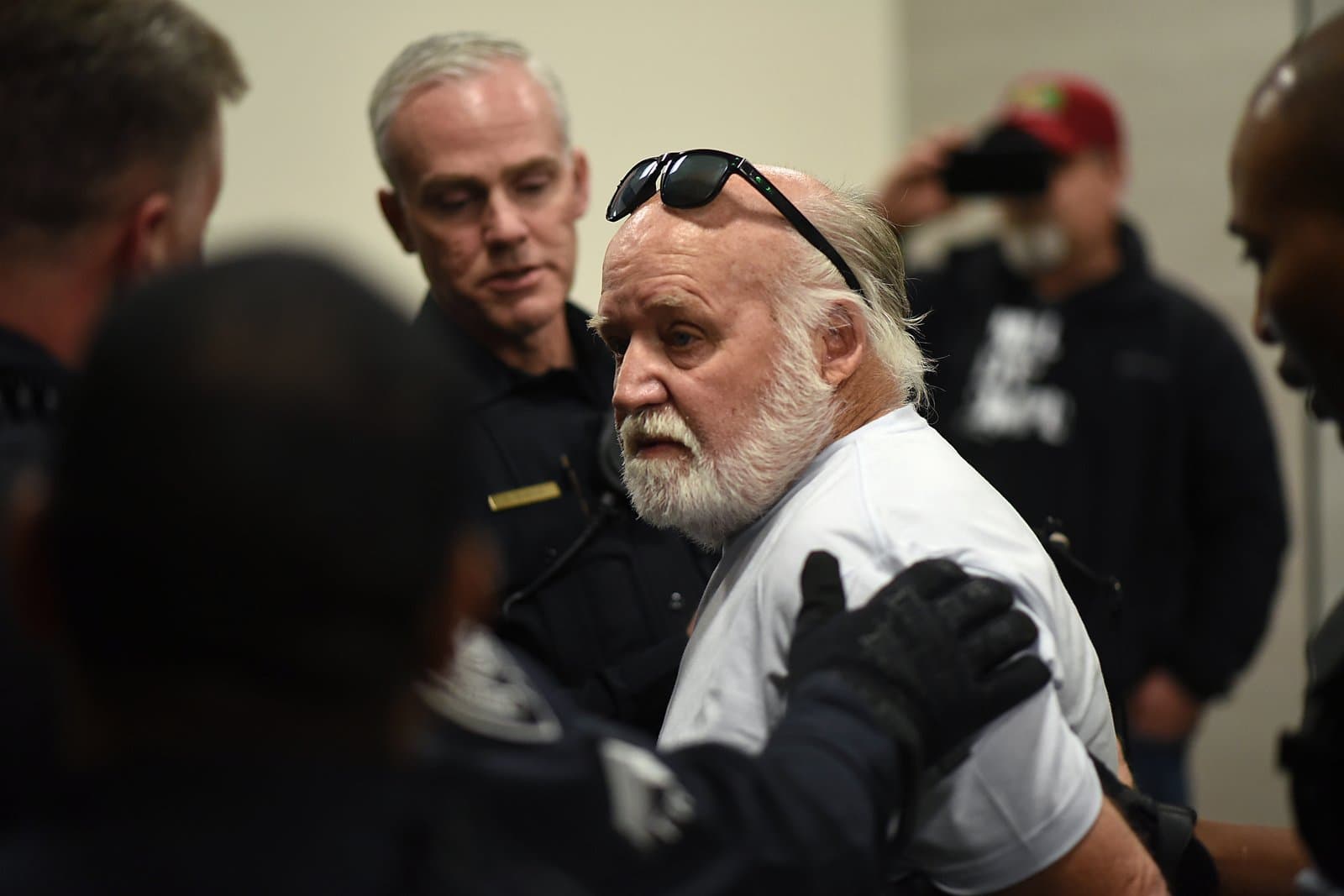 An older man with a white beard and sunglasses on his head is seen interacting with uniformed officers at an Anchorage Assembly meeting. Other individuals are visible in the background, suggesting a public gathering.