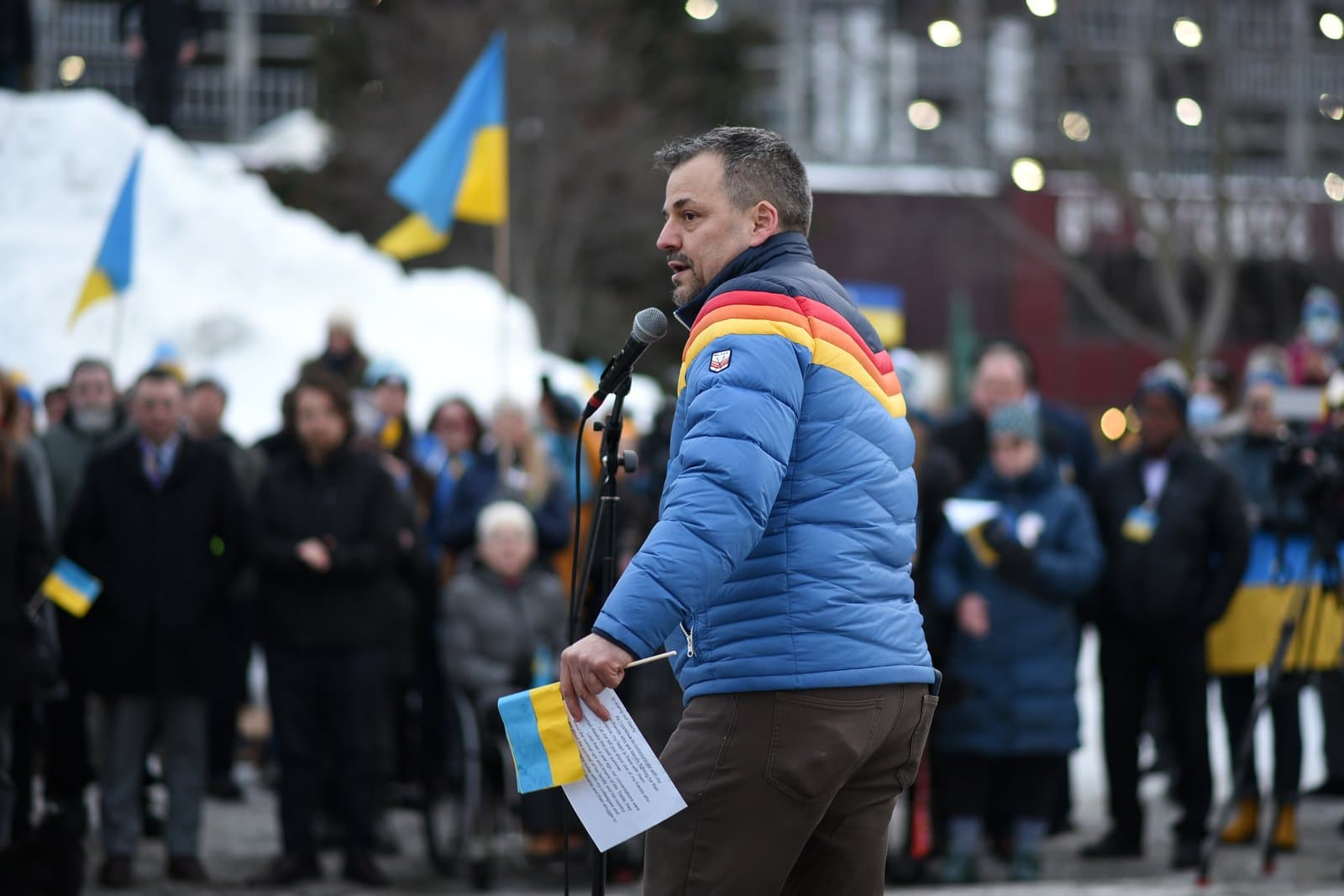 A man speaks into a microphone at an outdoor rally in Anchorage, holding a small Ukrainian flag and a printed paper.