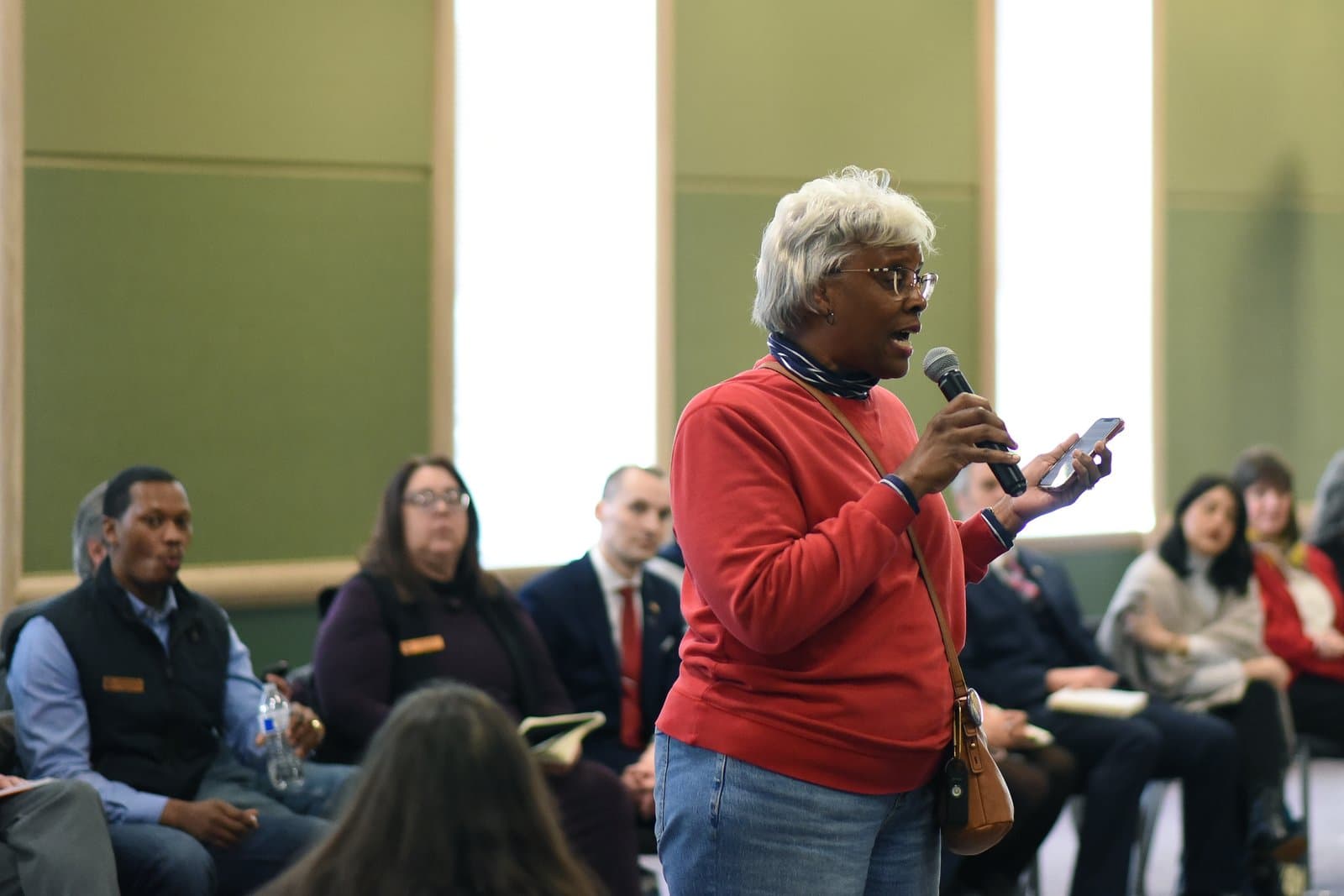 A woman speaks into a microphone while holding a smartphone during the 2024 Anchorage Legislative Town Hall meeting in Alaska.