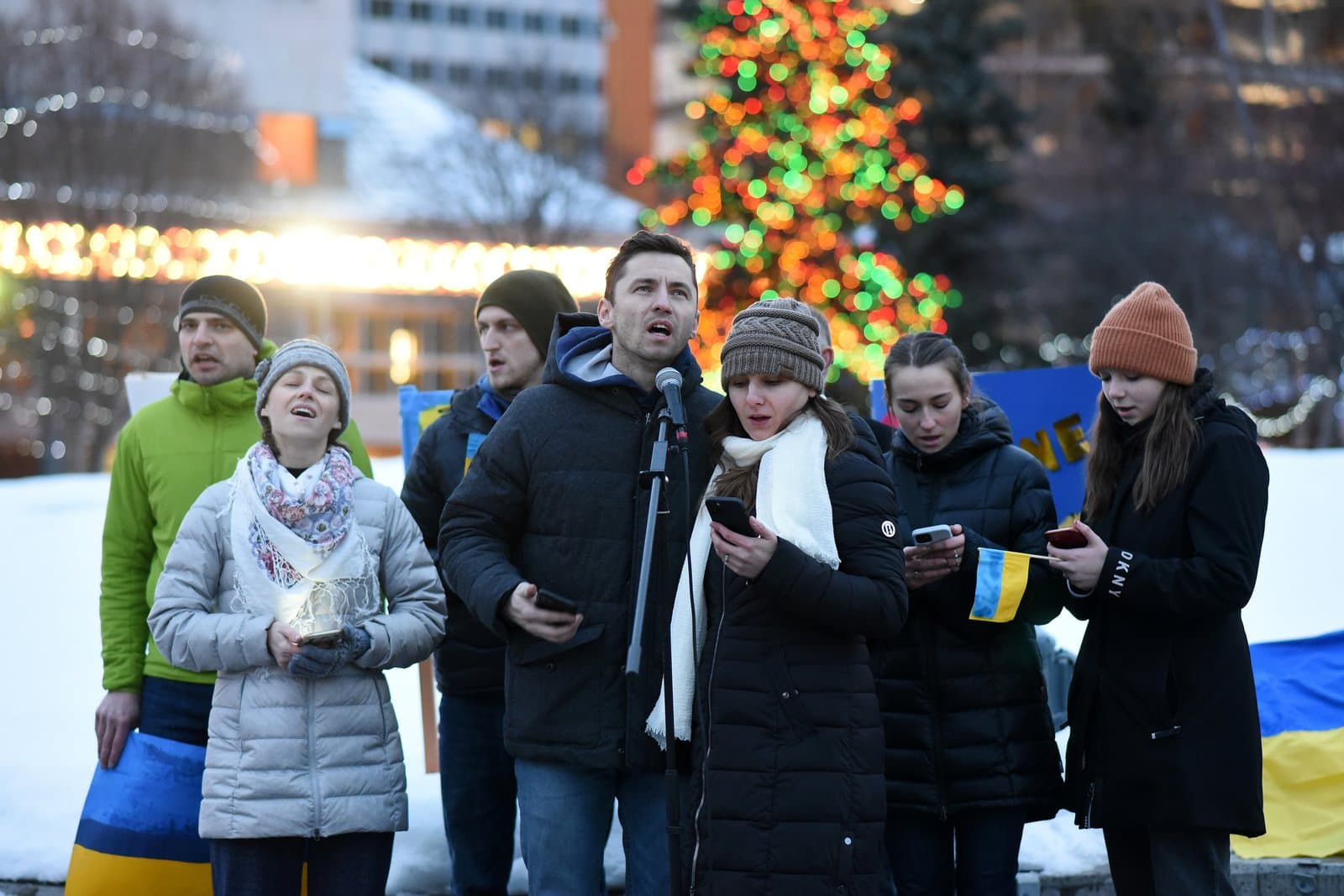 A group of people gather outdoors in Anchorage, Alaska, holding a Ukrainian flag and mobile devices during a winter rally for Ukraine.