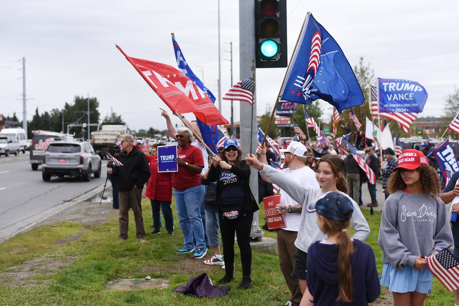 Supporters gather at a roadside rally in midtown Anchorage, holding campaign flags and signs along a busy street in August 2025.