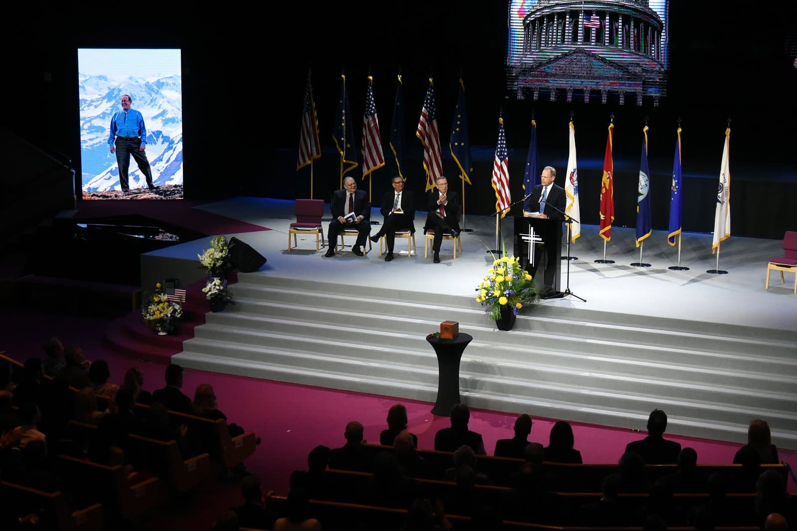 A speaker addresses attendees at the memorial service for Don Young, held in a large auditorium featuring American and state flags.