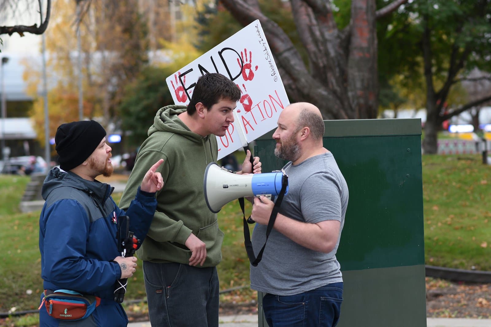 Three individuals engage in a public protest in Anchorage, with one person holding a sign reading End Occupation and another using a megaphone.