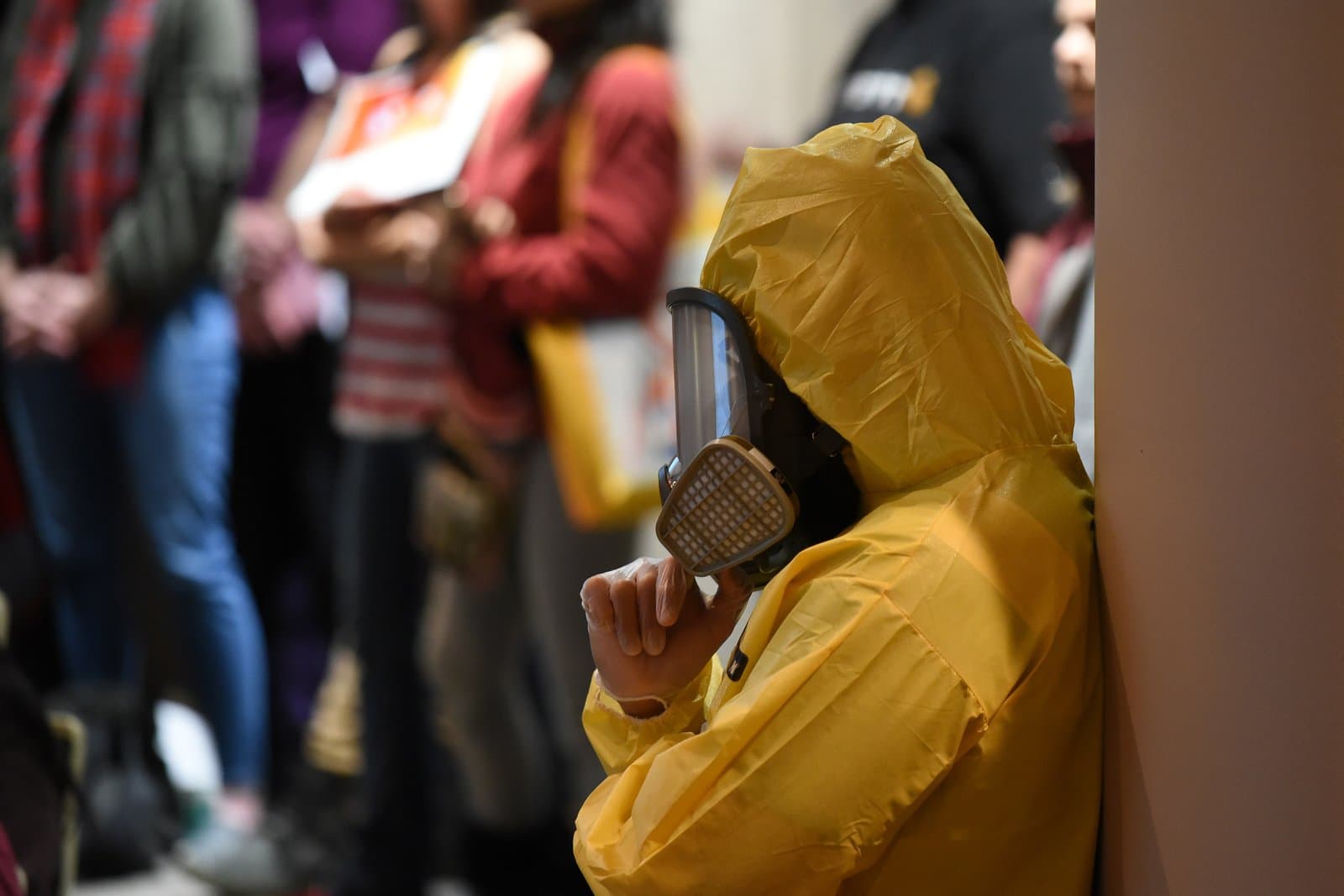 A person in a bright yellow hazmat suit and gas mask leans against a wall, with blurred figures of other attendees in the background at an Anchorage Assembly meeting.