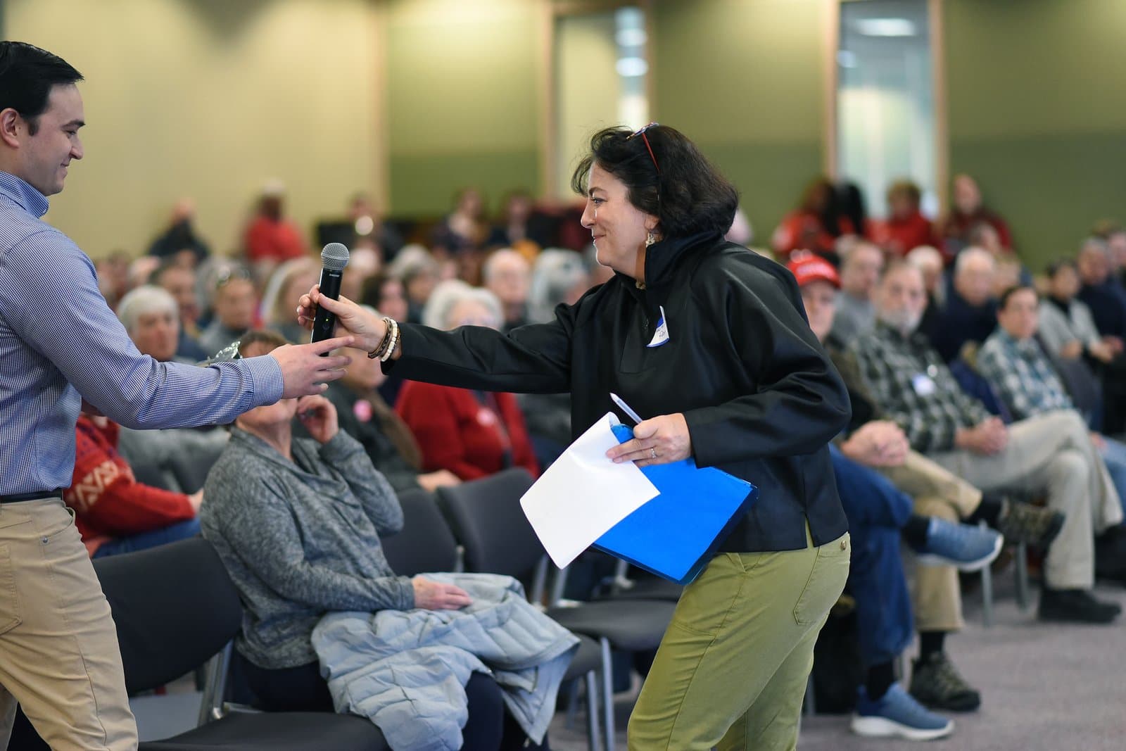 A woman hands a microphone to a man during the 2024 Anchorage Legislative Town Hall meeting held in an indoor community space.