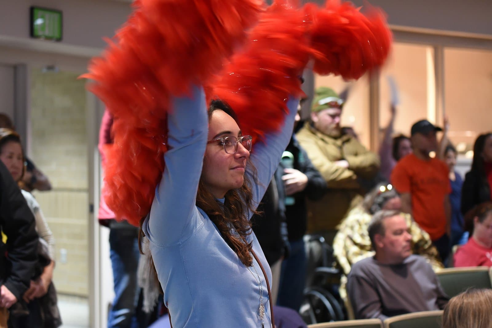 A woman with glasses and long brown hair raises two large, red, feathery objects above her head, smiling slightly, while attending an Anchorage Assembly meeting. Other people are visible in the blurred background, some also appearing to be engaged in the event.