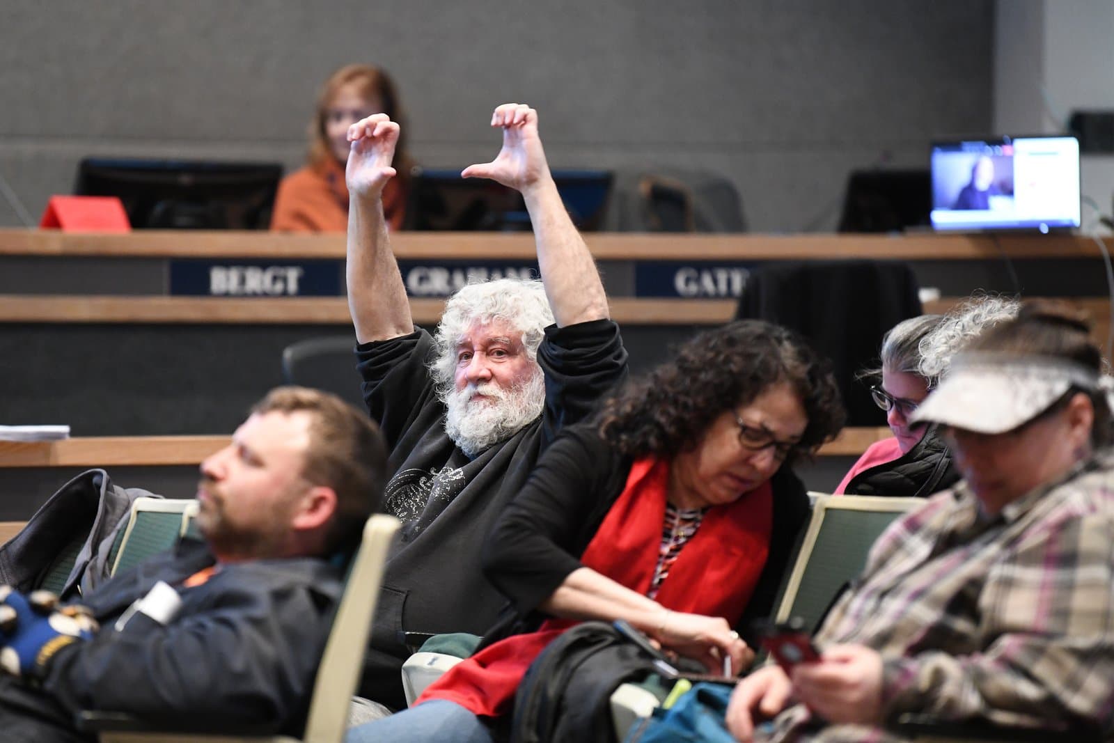 A man with a white beard and dark hoodie raises both hands in a public meeting, looking towards the camera. Other attendees are seated around him, some looking down at their phones. In the background, signs read 'BERGT', 'GRAHAM', and 'GATE', and a screen displays a video call.