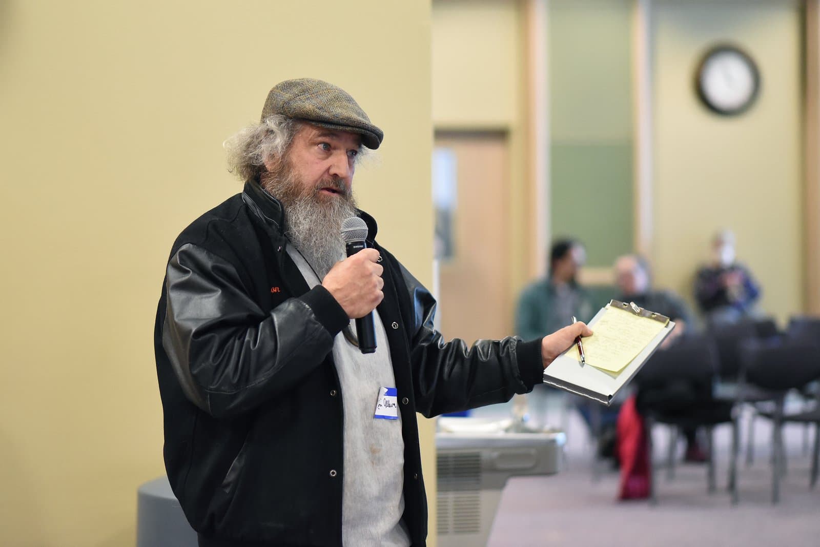 A man with a beard and flat cap speaks into a microphone while holding a clipboard at the 2024 Anchorage Legislative Town Hall.