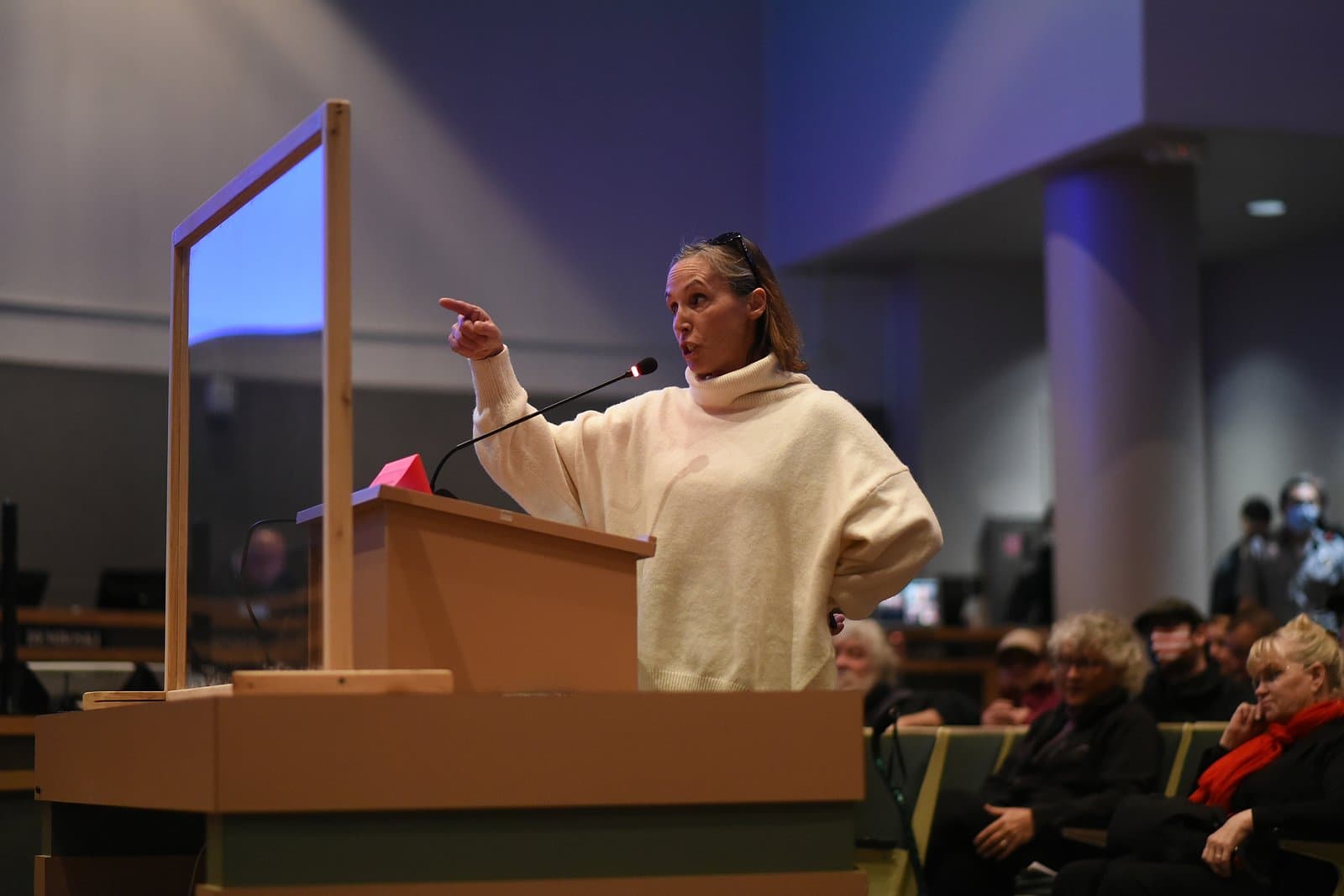A woman in a cream-colored turtleneck sweater speaks passionately at a wooden podium with a microphone, pointing her right hand, during an Anchorage Assembly meeting. A clear barrier stands to her left, and other attendees are visible in the background.