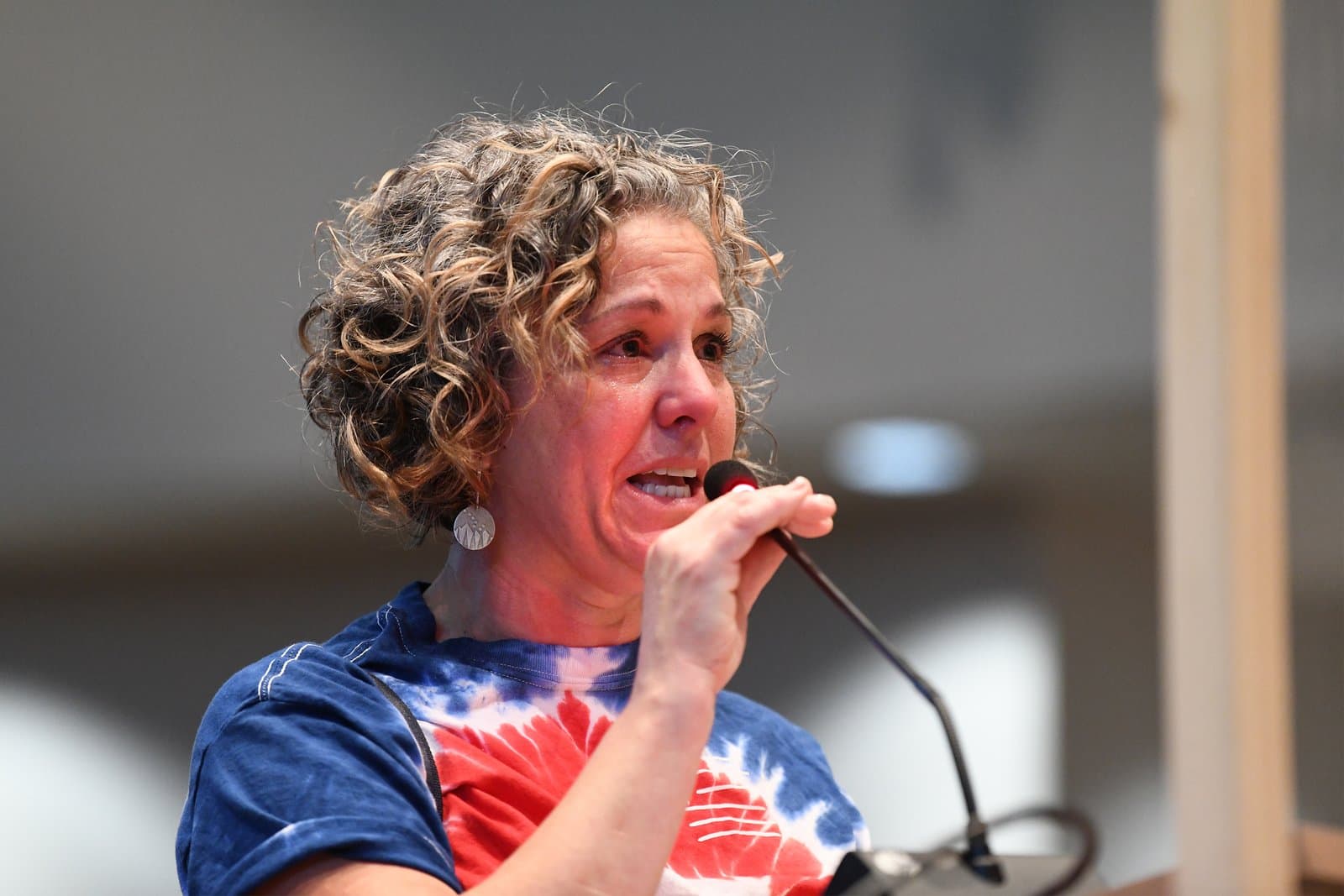 A woman with curly hair and a red, white, and blue tie-dye shirt speaks emotionally into a microphone at an Anchorage Assembly Meeting, her face showing distress.