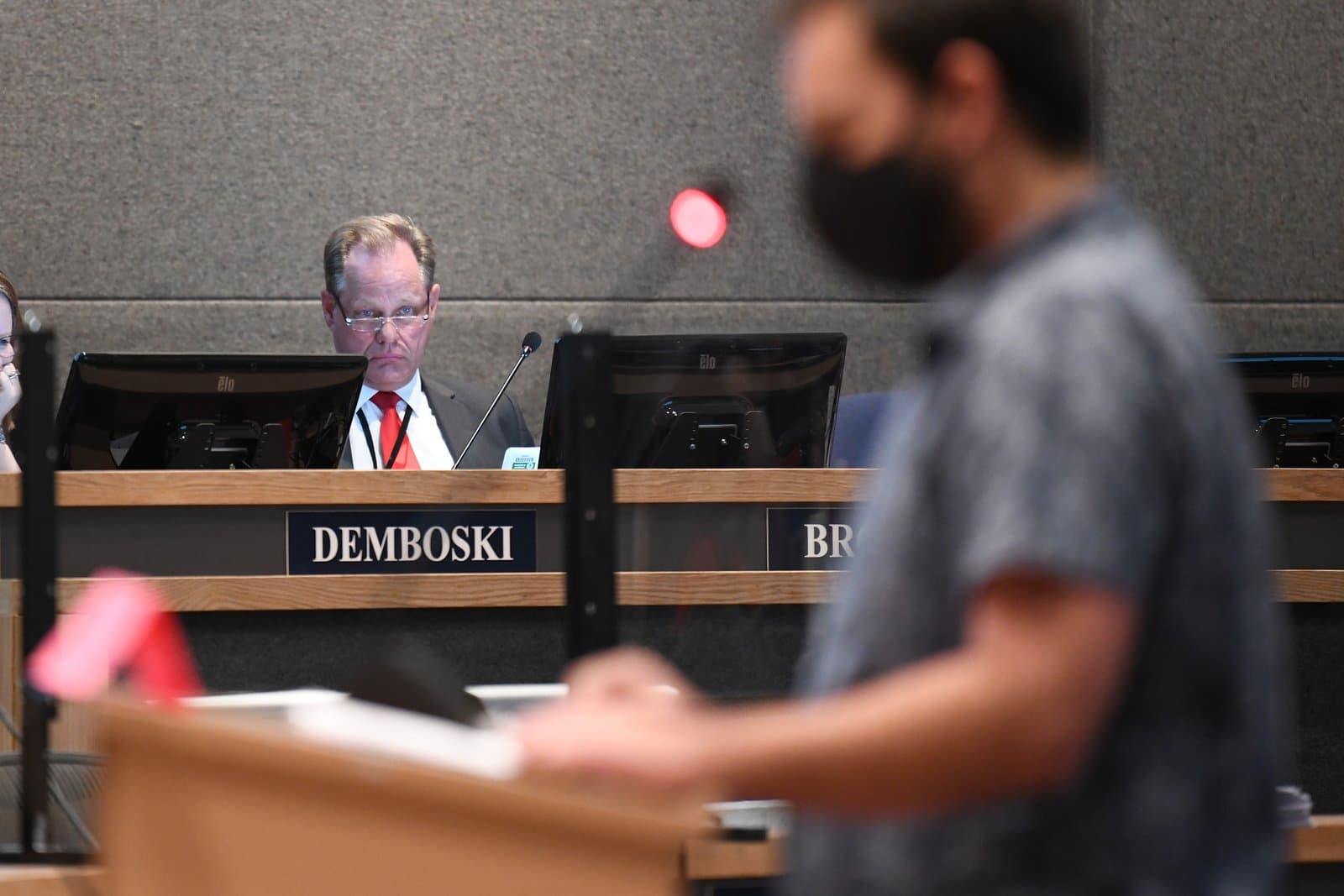 A man identified as Demboski, wearing a suit and red tie, sits behind a desk with monitors during an Anchorage Assembly meeting. In the blurred foreground, another individual wearing a face mask stands at a podium.