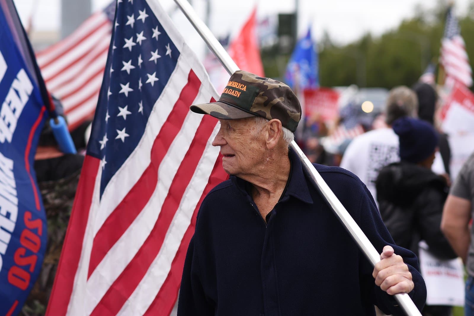 An elderly man wearing a camouflage Make America Great Again hat holds an American flag during a pro-Trump rally in midtown Anchorage, Alaska.