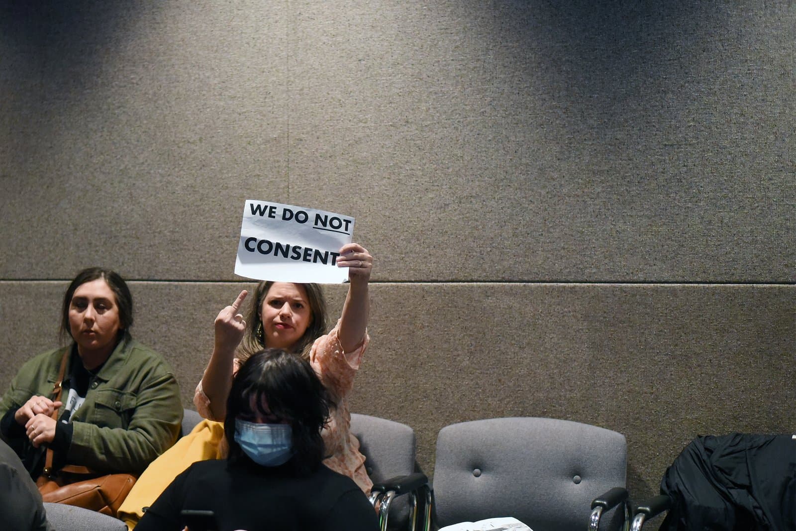 A woman holds a sign reading We Do Not Consent while gesturing with her middle finger during an Anchorage Assembly meeting in October 2021.