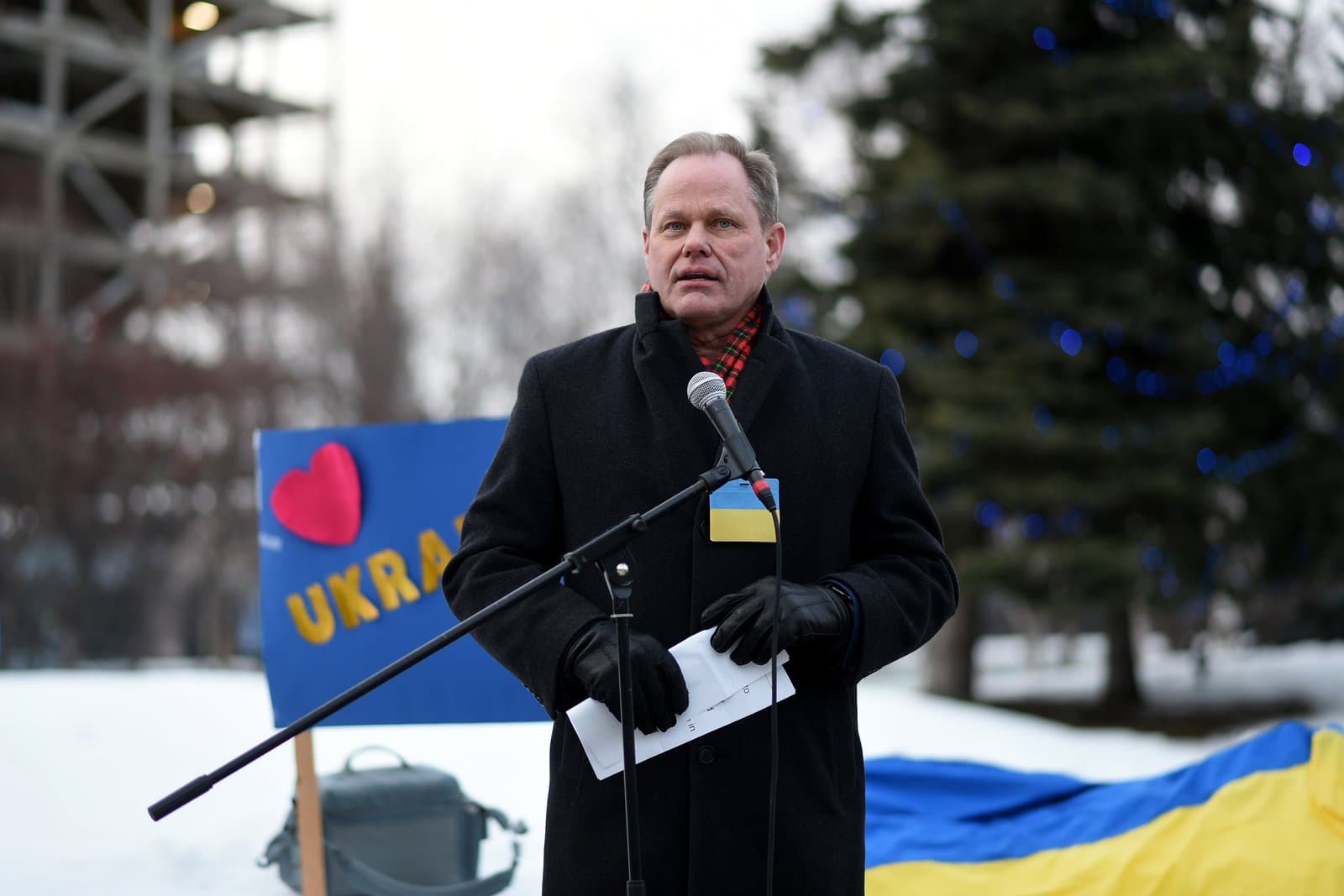 A man wearing a black coat and a Ukrainian flag pin speaks into a microphone at an outdoor rally in Anchorage, Alaska.