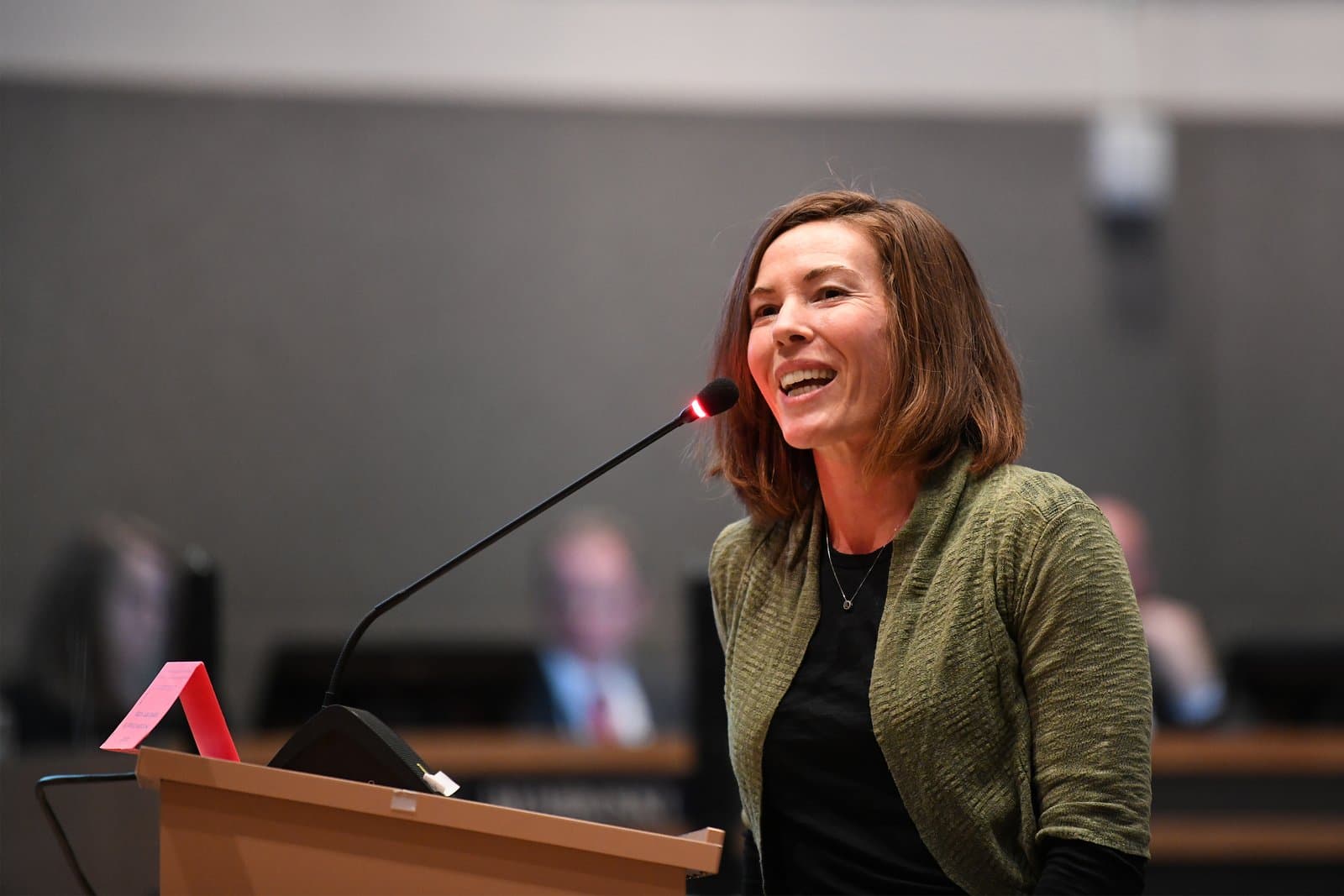 A woman with brown hair, wearing a green cardigan and black shirt, smiles while speaking into a microphone at a wooden podium during an indoor meeting.