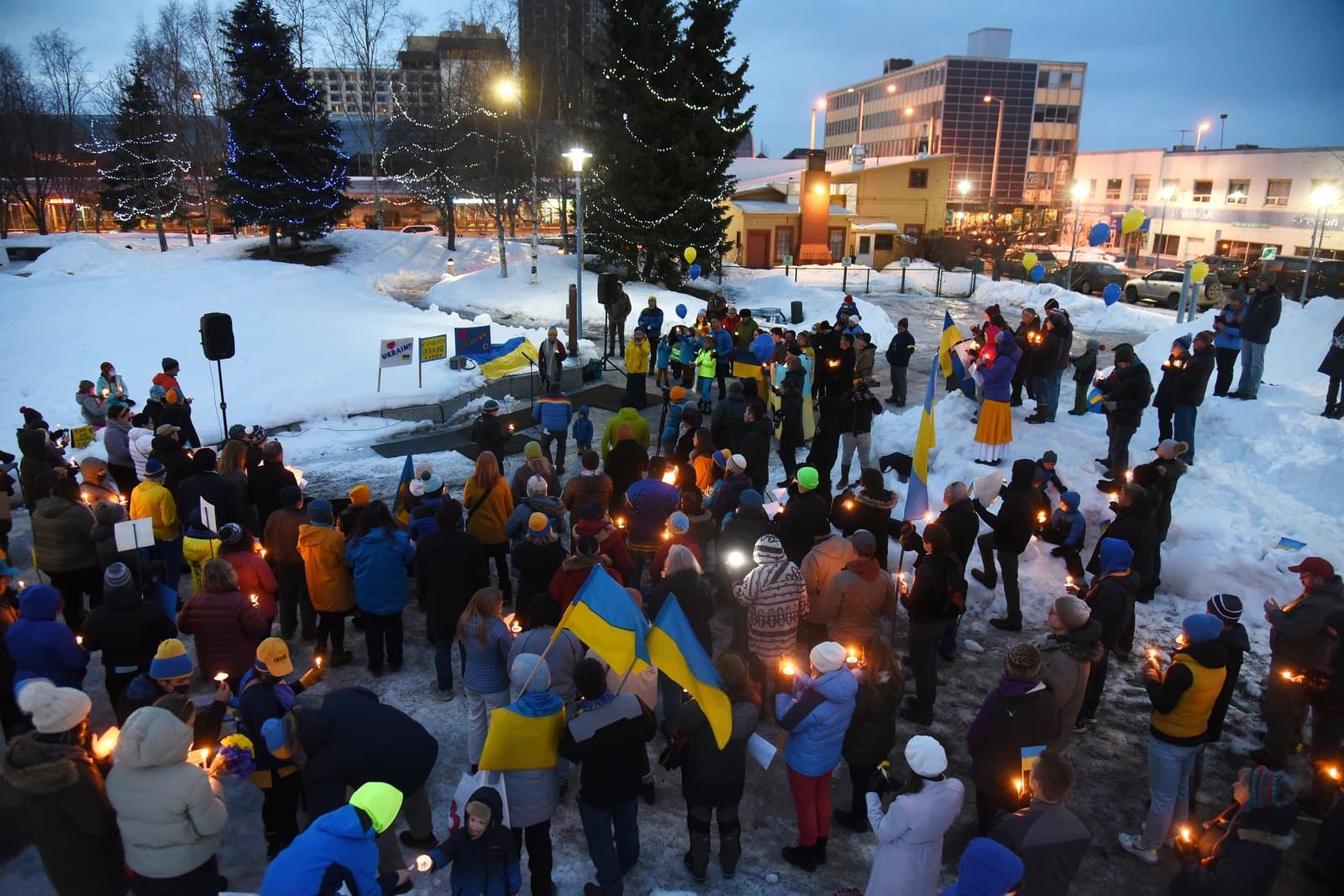 A crowd gathers in a snow-covered Anchorage park for a candlelight vigil in support of Ukraine, featuring blue and yellow flags.