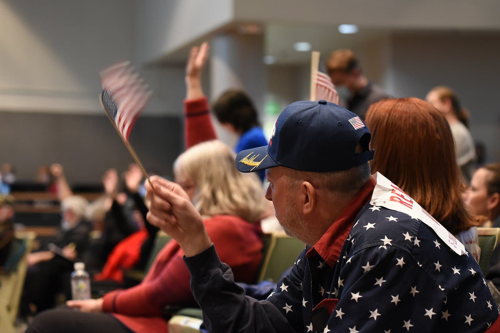 A man in a star-spangled shirt and a blue cap holds a small American flag at an Anchorage Assembly meeting, with other attendees visible in the background, some also holding flags or raising their hands.