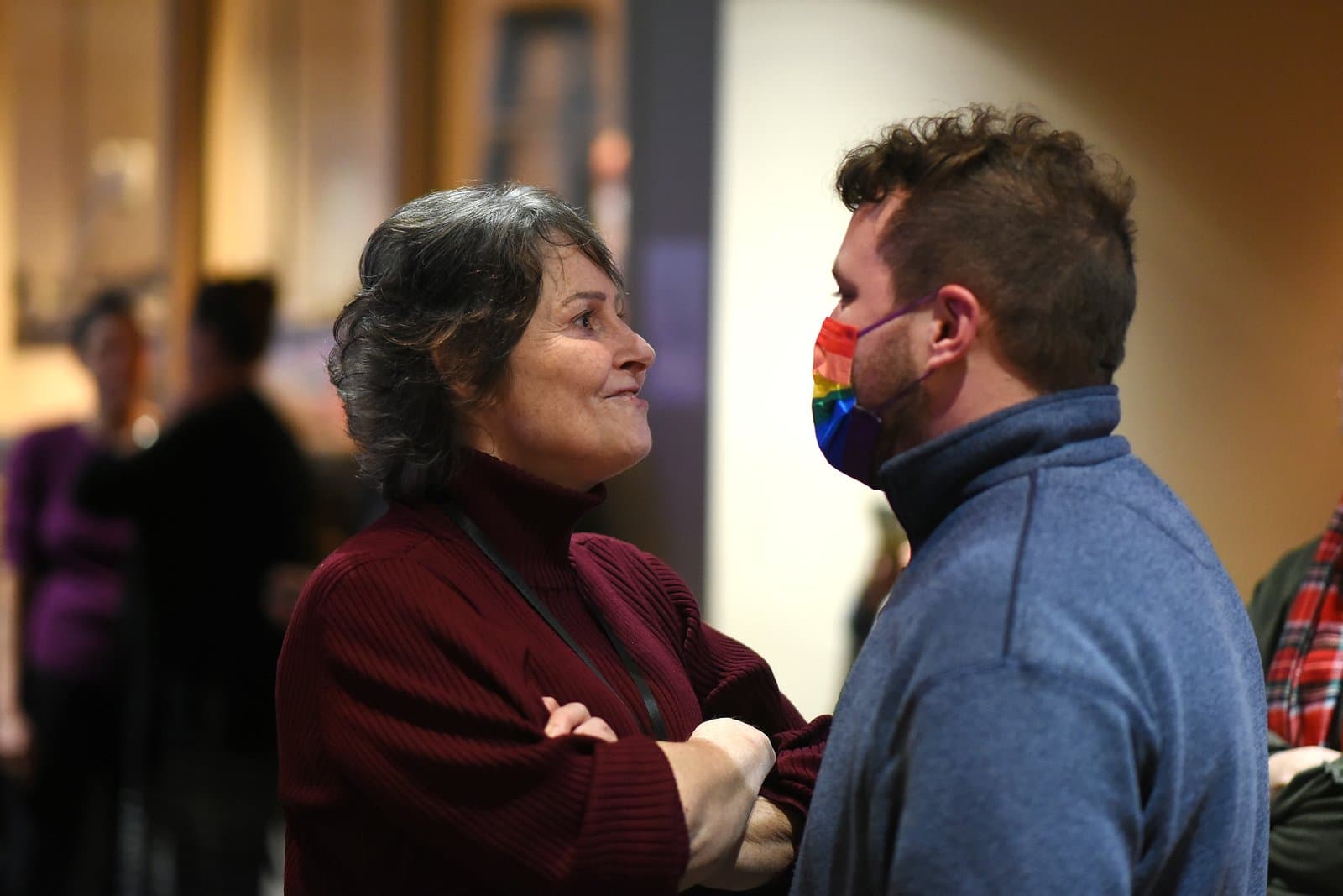 An older woman with dark, curly hair and a dark red turtleneck smiles while looking at a younger man wearing a blue fleece and a rainbow-colored face mask, both engaged in conversation at an indoor meeting.
