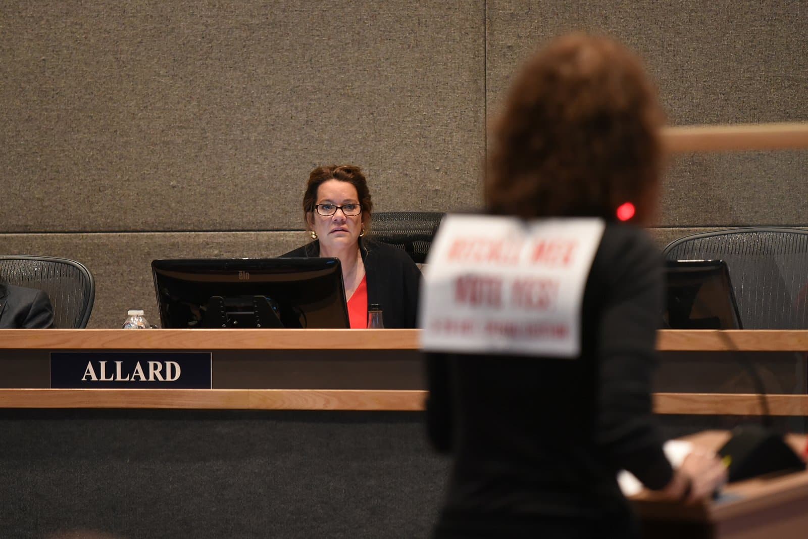 A woman identified as Assembly Member Allard, wearing glasses and a red top under a black jacket, sits at a desk in an assembly chamber, looking intently towards an out-of-focus person in the foreground. A monitor with 'elo' branding is on the desk.