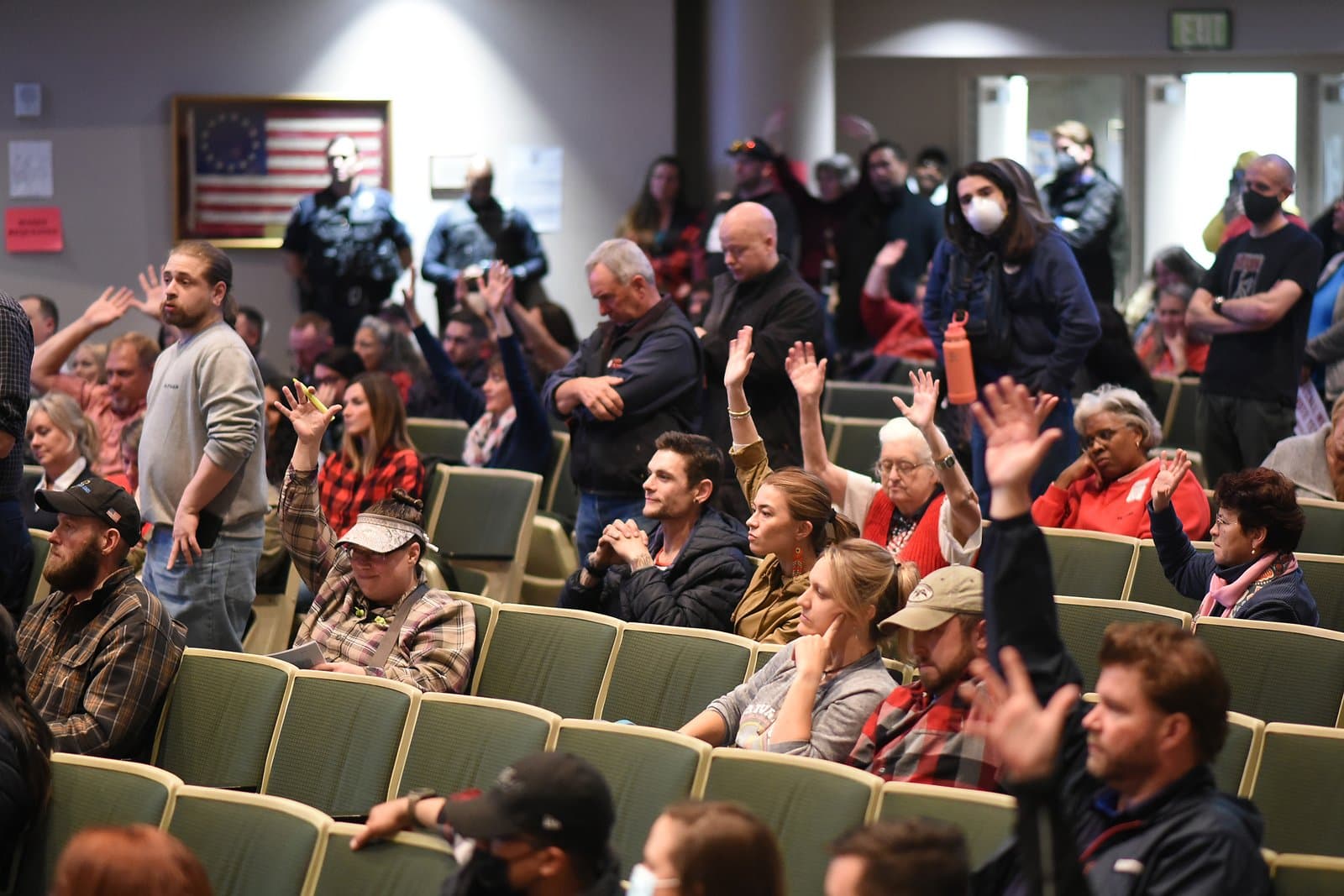 A diverse group of people, including men and women of various ages, are seated in an auditorium, with many raising their hands in what appears to be a vote or to ask a question during an Anchorage Assembly meeting. Police officers stand in the background, and a historic American flag hangs on the wall.