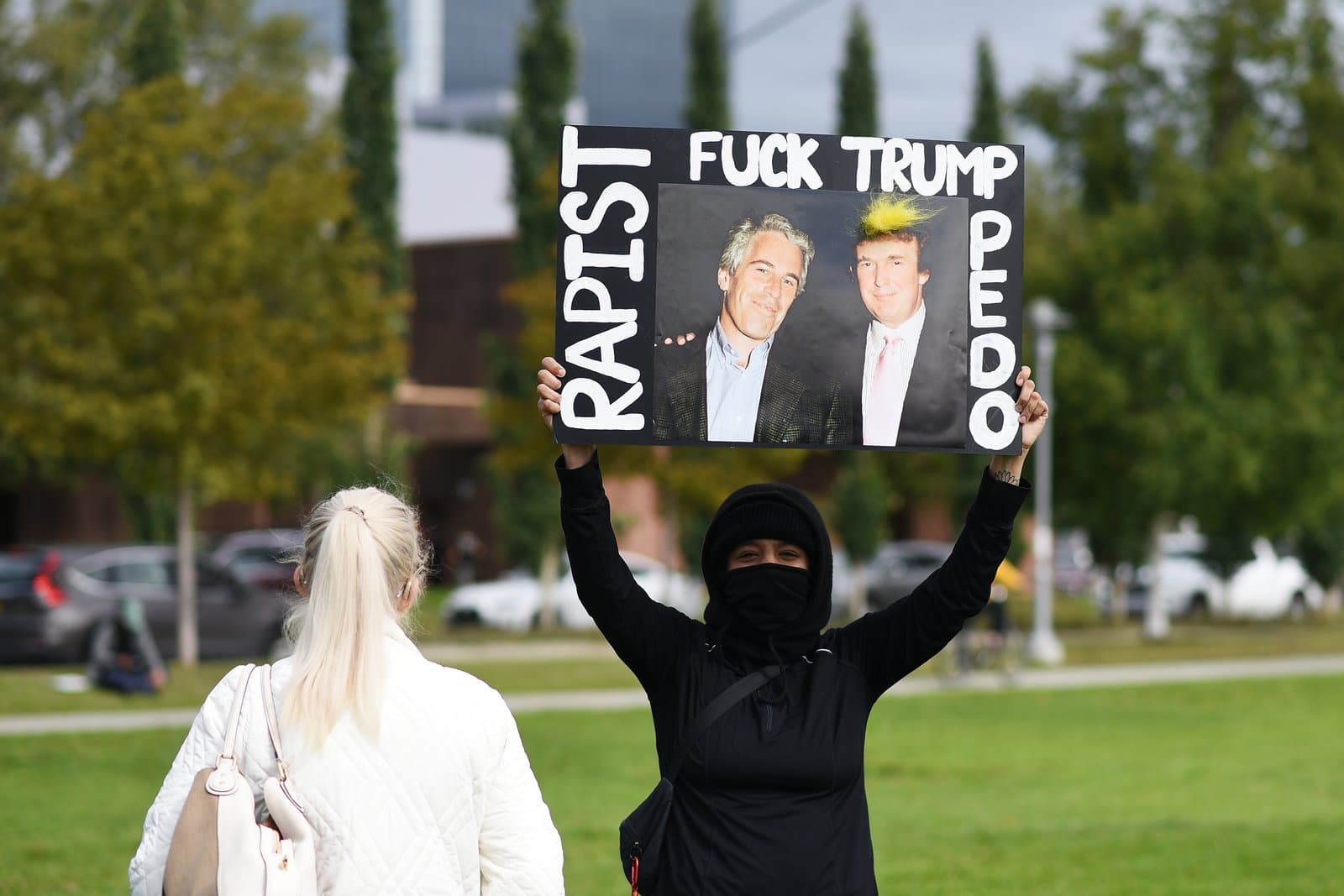 A masked protester holds a sign featuring Jeffrey Epstein and Donald Trump during a demonstration in downtown Anchorage, Alaska.