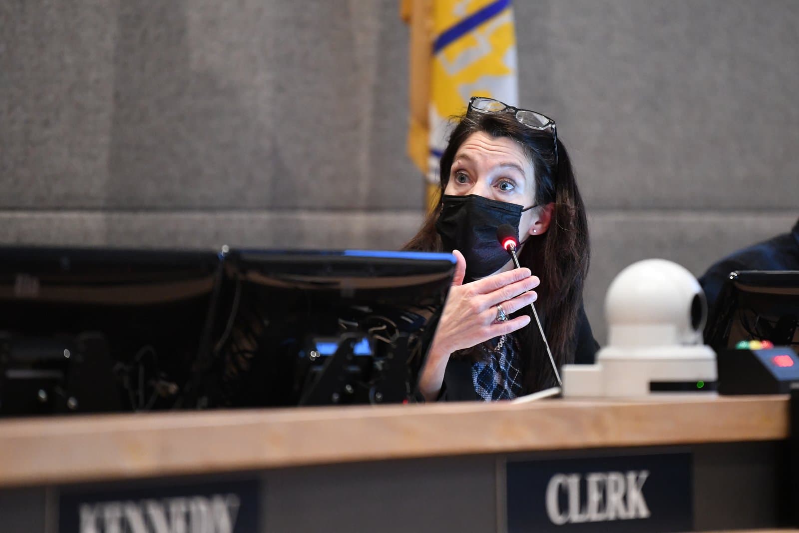 A woman wearing a black face mask and glasses speaks animatedly into a microphone at an Anchorage Assembly meeting, with monitors and a 'CLERK' sign visible on the table.