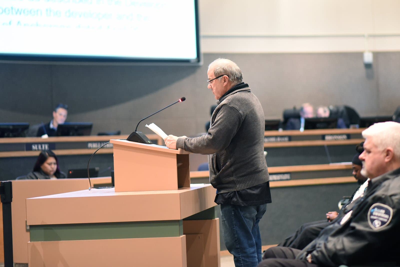 David Nyman stands at a podium reading a document while testifying before the Anchorage Assembly in a municipal meeting room.
