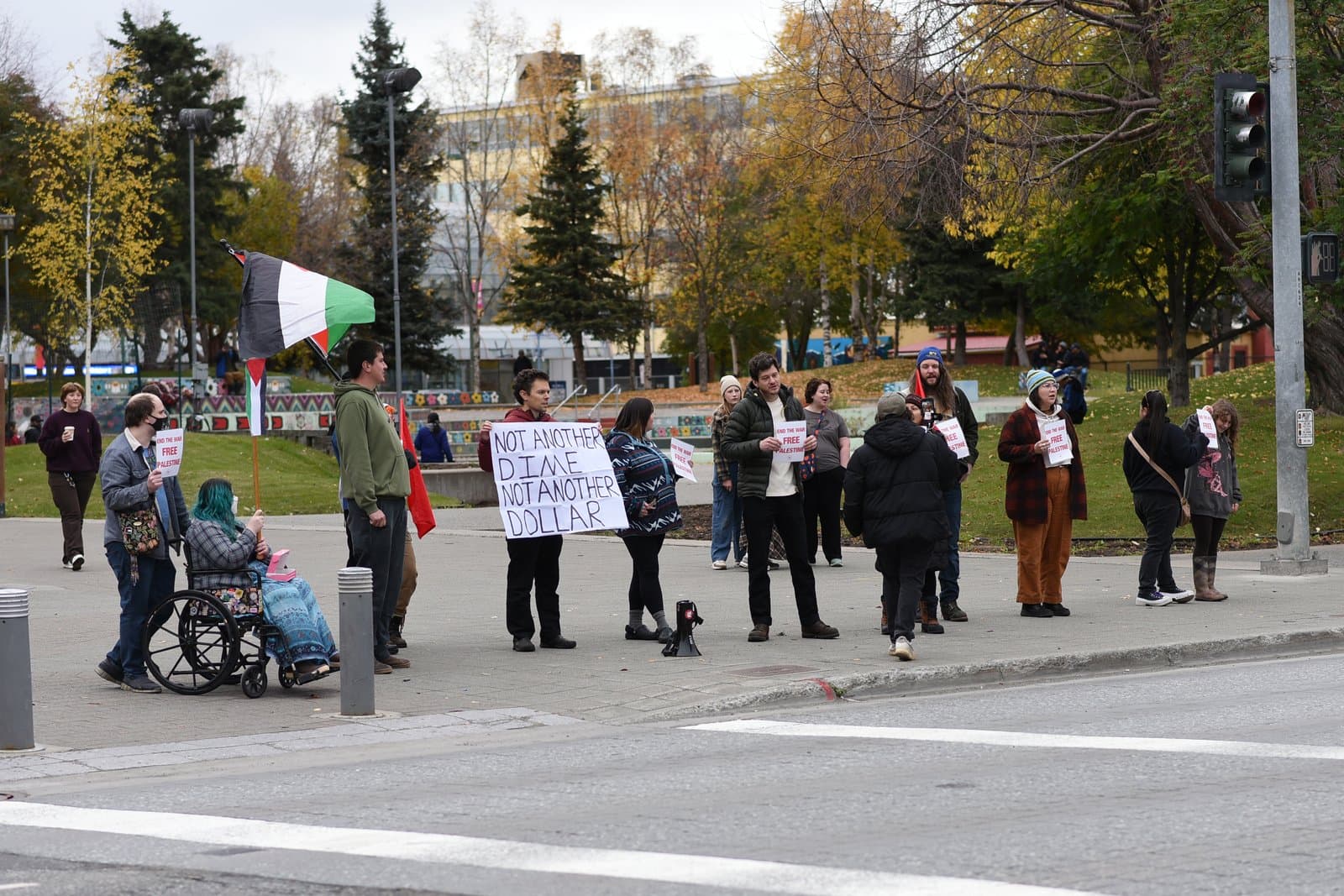 A group of protesters holding signs and a Palestinian flag stands at an intersection in downtown Anchorage during an autumn afternoon.