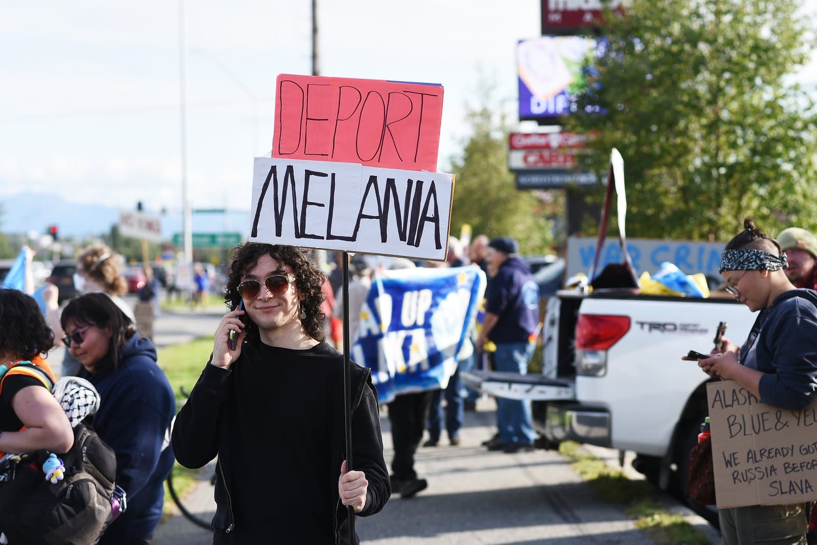 A young person holds a protest sign reading "Deport Melania" while talking on a cell phone during a rally in midtown Anchorage, Alaska.