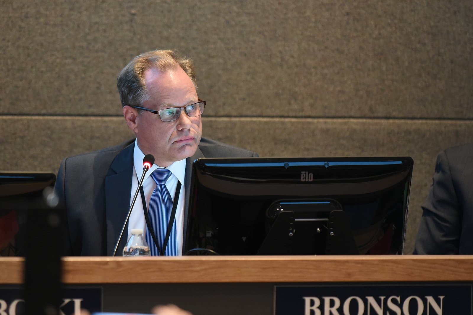 A man in a suit and glasses, identified by a 'Bronson' nameplate, sits intently at a table with a microphone and computer monitor during an Anchorage Assembly meeting.