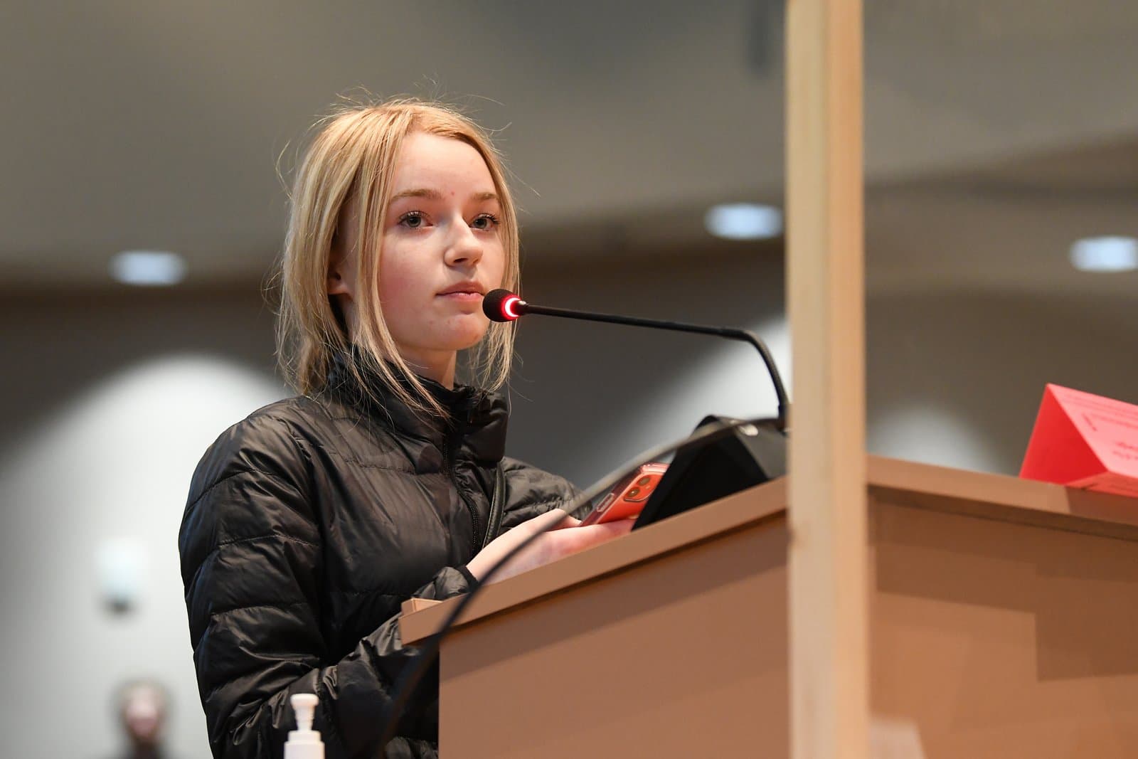 A young blonde woman in a black puffer jacket stands at a wooden podium, speaking into a microphone with a red light, during an Anchorage Assembly meeting.