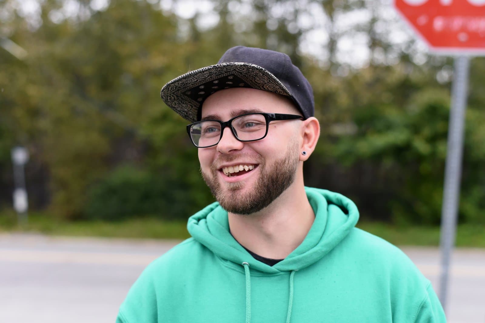 A smiling man wearing a black baseball cap and glasses stands outdoors in front of a blurred background with a stop sign.