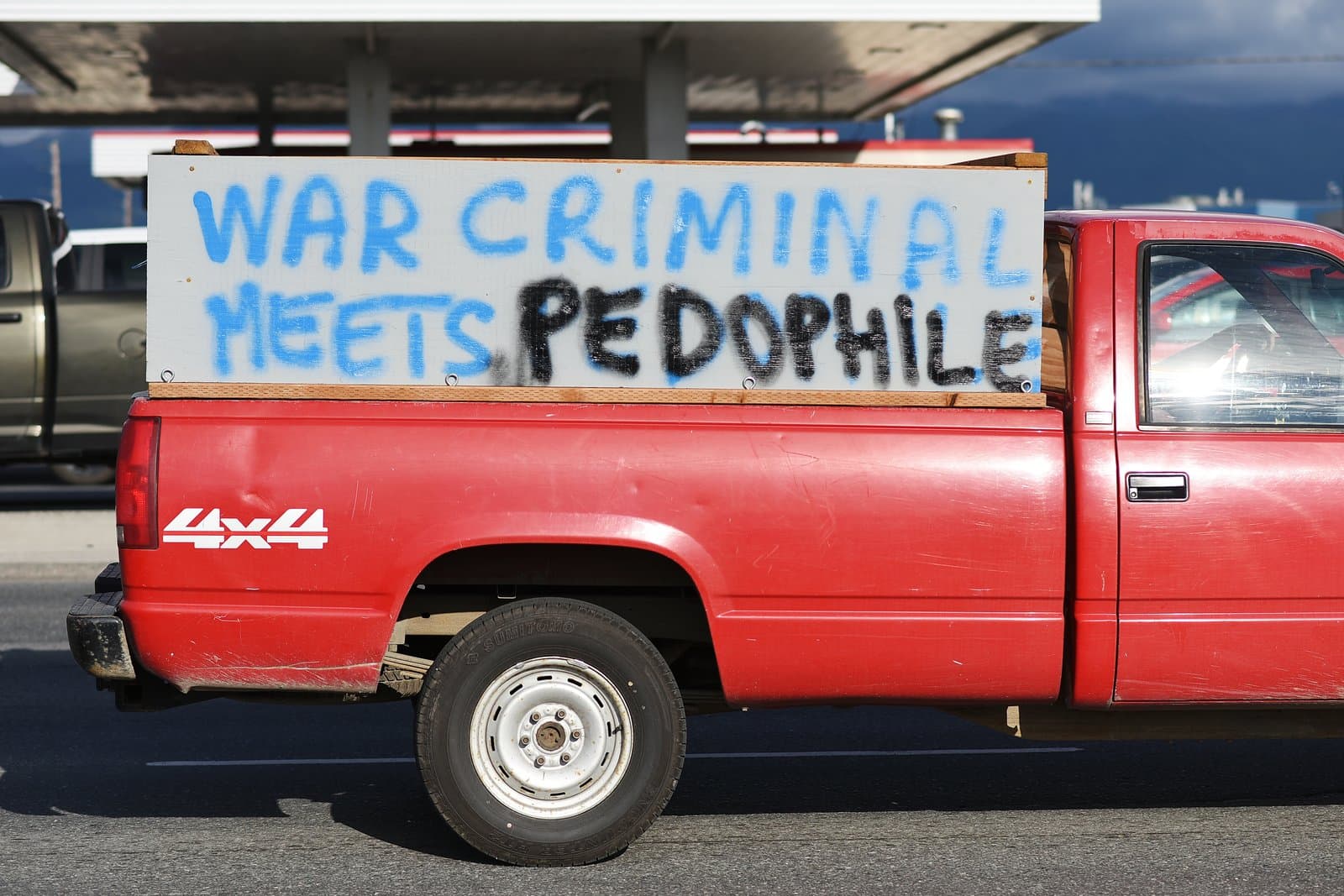 A red pickup truck displays a hand-painted sign reading War Criminal Meets Pedophile during a protest in midtown Anchorage, Alaska.