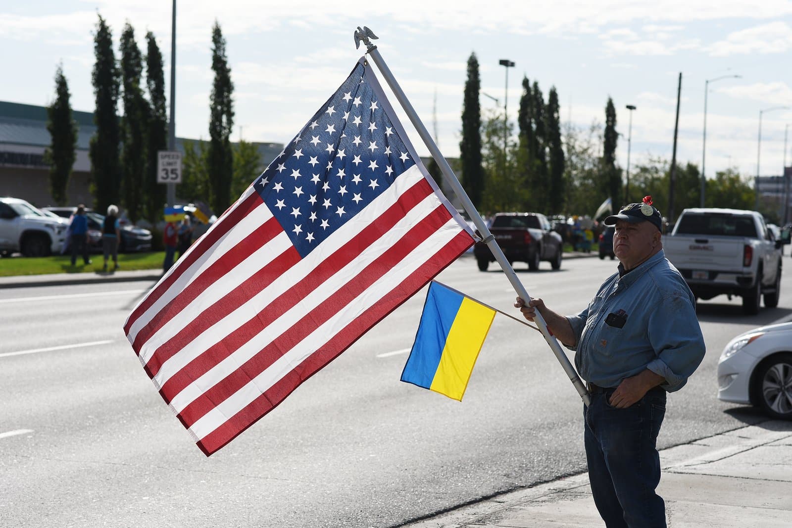 A man stands on a sidewalk in midtown Anchorage holding an American flag and a smaller Ukrainian flag during a public protest.