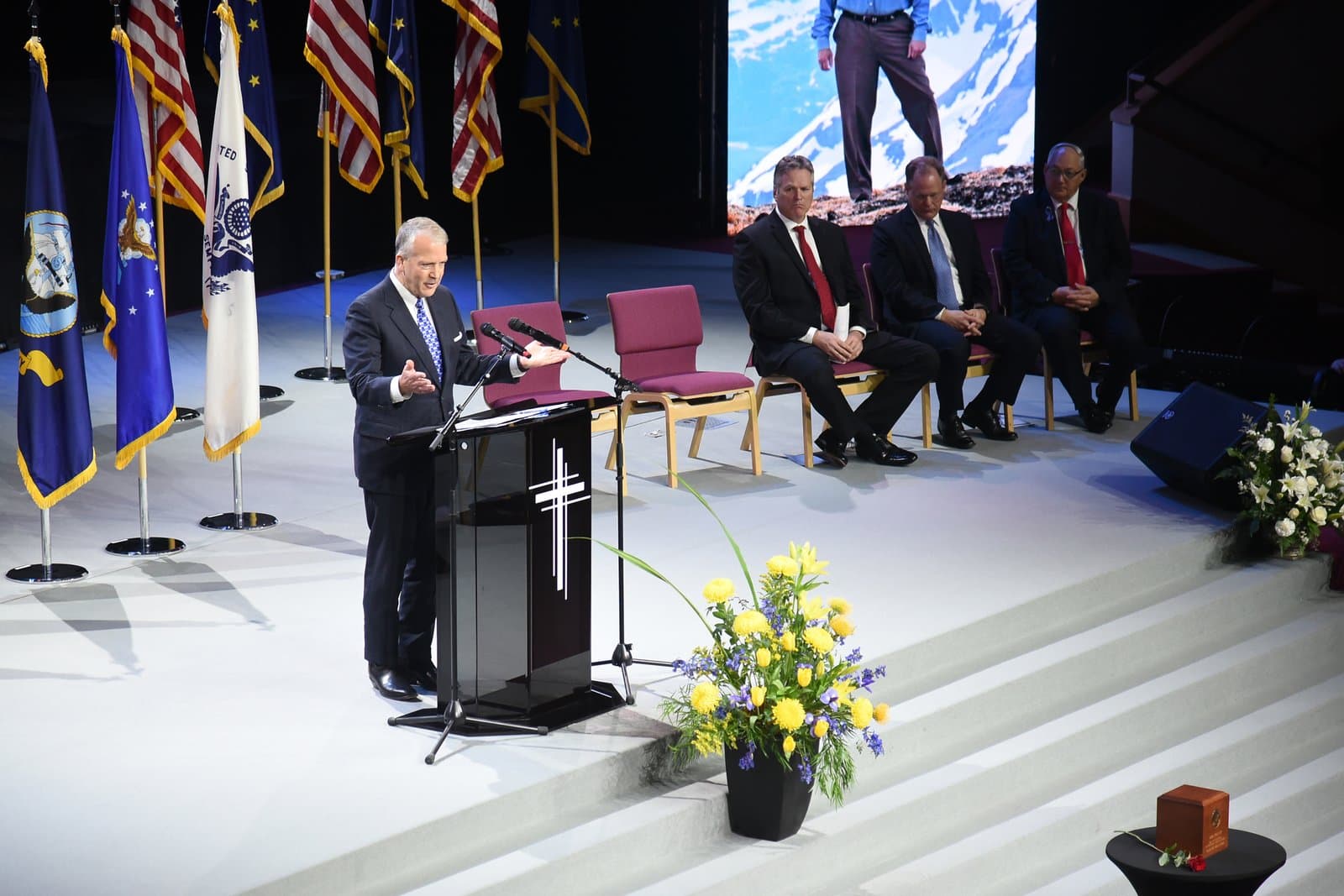 A speaker addresses attendees at the memorial service for Don Young, held in a formal auditorium with American and state flags displayed.