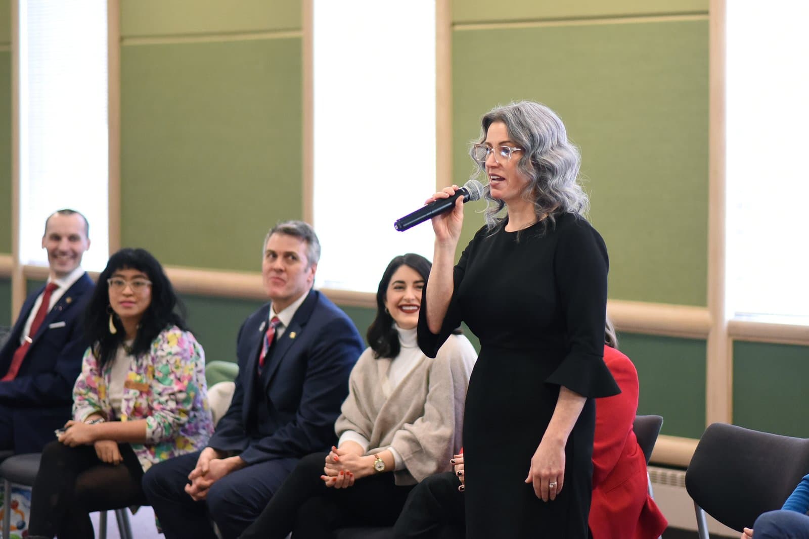A woman speaks into a microphone at the 2024 Anchorage Legislative Town Hall while seated officials listen in the background.