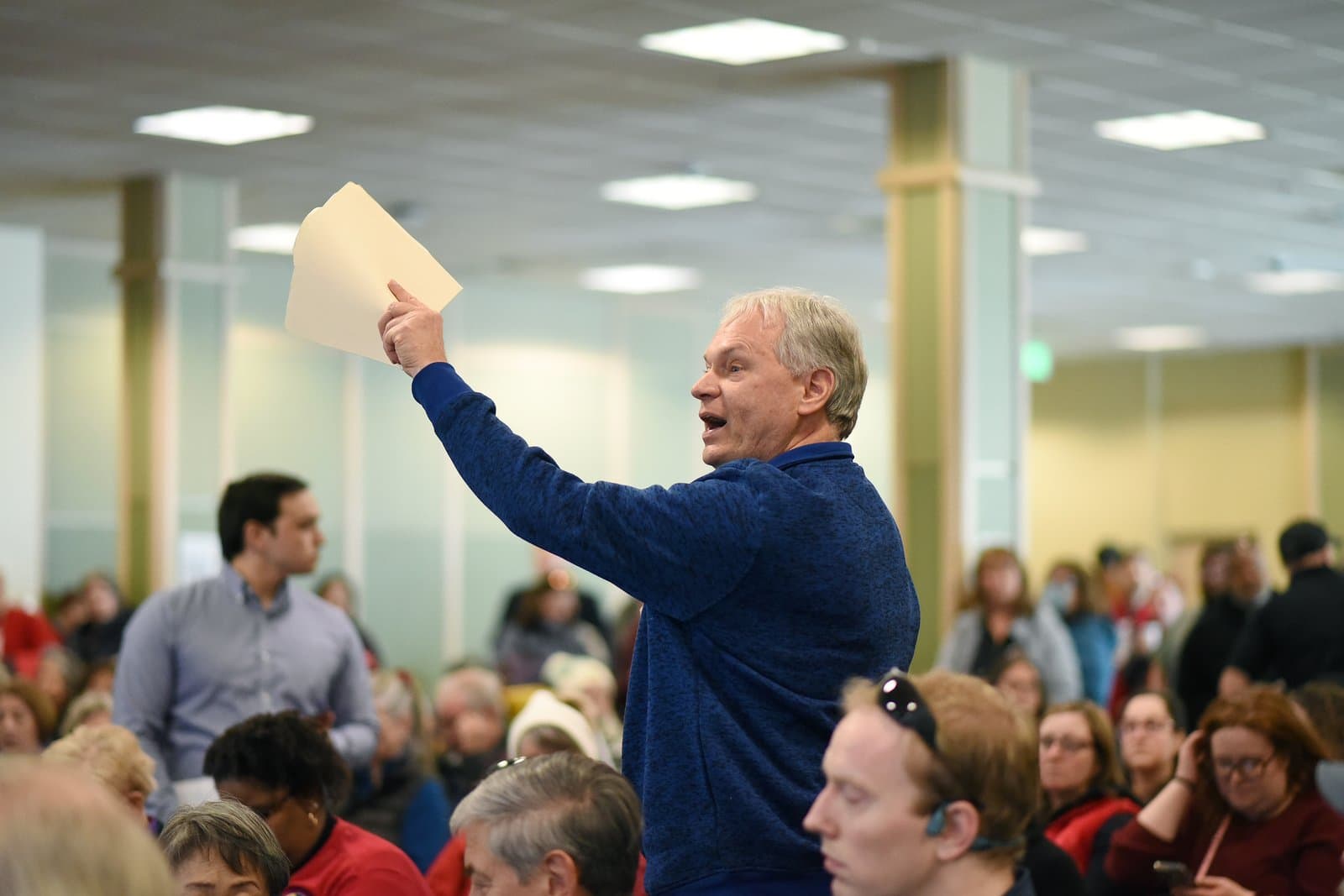 A man raises a document while speaking during the 2024 Anchorage Legislative Town Hall held in an indoor community space.