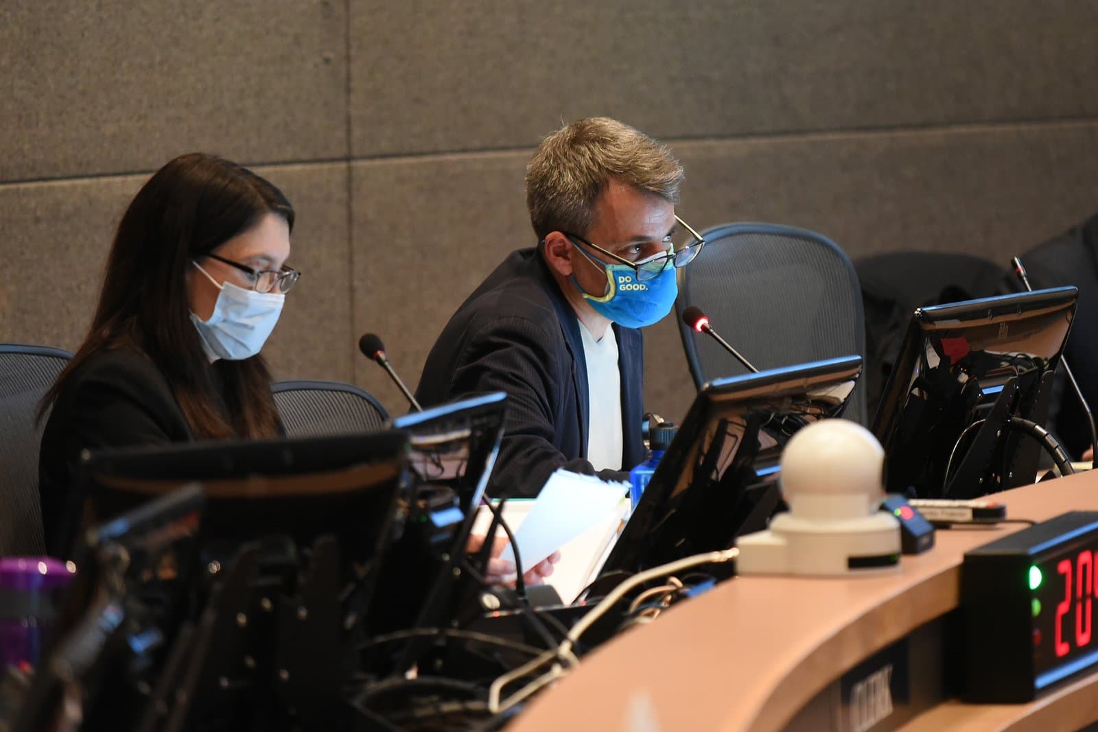 Two masked individuals, an Asian woman and a Caucasian man, are seated at a table with monitors and microphones, participating in an Anchorage Assembly meeting. The man's blue mask reads 'DO GOOD.' in yellow text, and a digital timer on the table shows '2:00'.