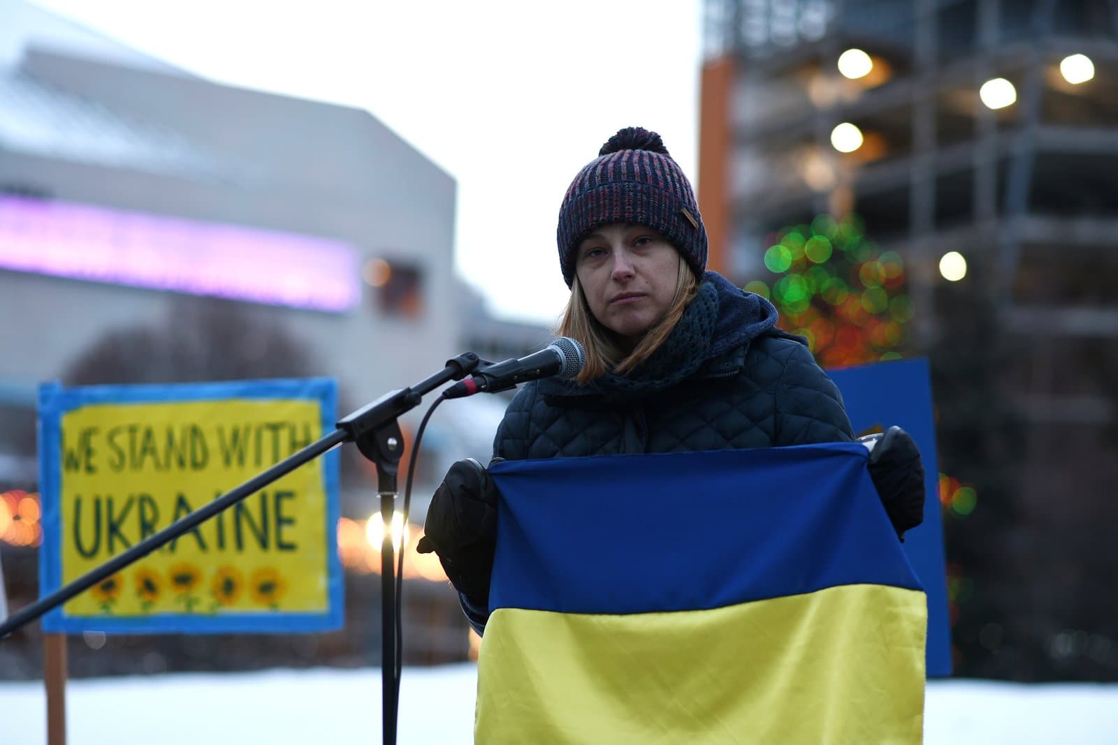 A woman wearing a winter hat and scarf holds a Ukrainian flag while speaking at an Anchorage rally supporting Ukraine.
