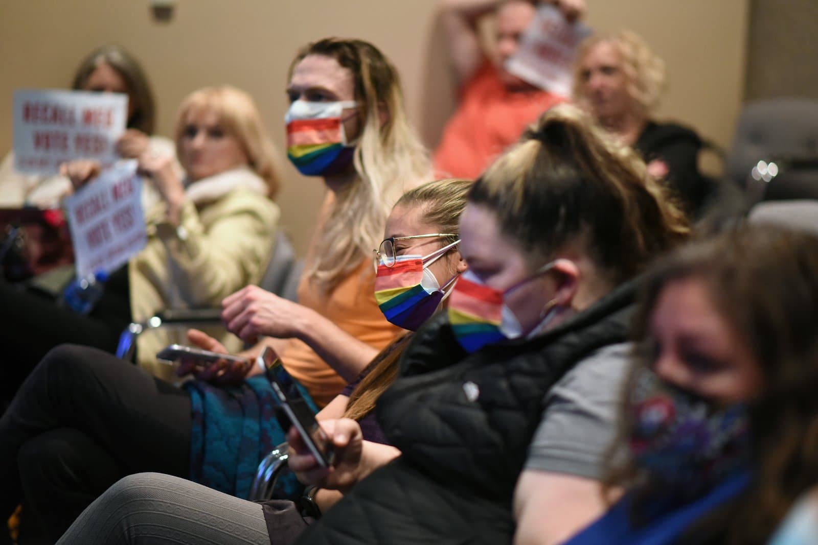 Attendees, some wearing rainbow-striped face masks, listen and hold signs during an Anchorage Assembly meeting, with one sign partially visible reading 'RECALL MICE VOTE YES!'.
