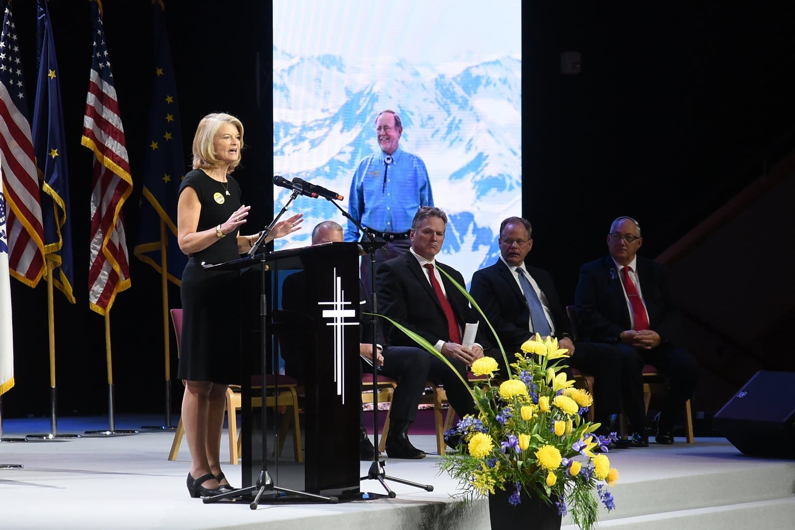 A woman speaks at a podium during the memorial service for Don Young, with a large mountain photograph displayed on the screen behind.