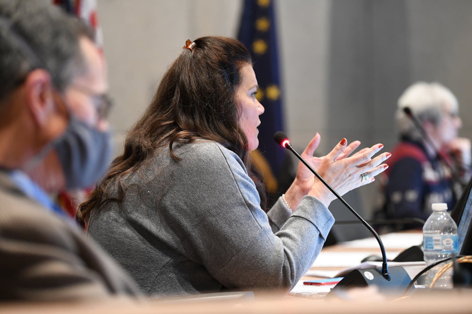 A woman with long dark hair, wearing a gray jacket, speaks into a microphone with a red light, gesturing with her hands. She is seated at a table in what appears to be a public meeting, with other blurred figures and an Alaskan flag in the background.