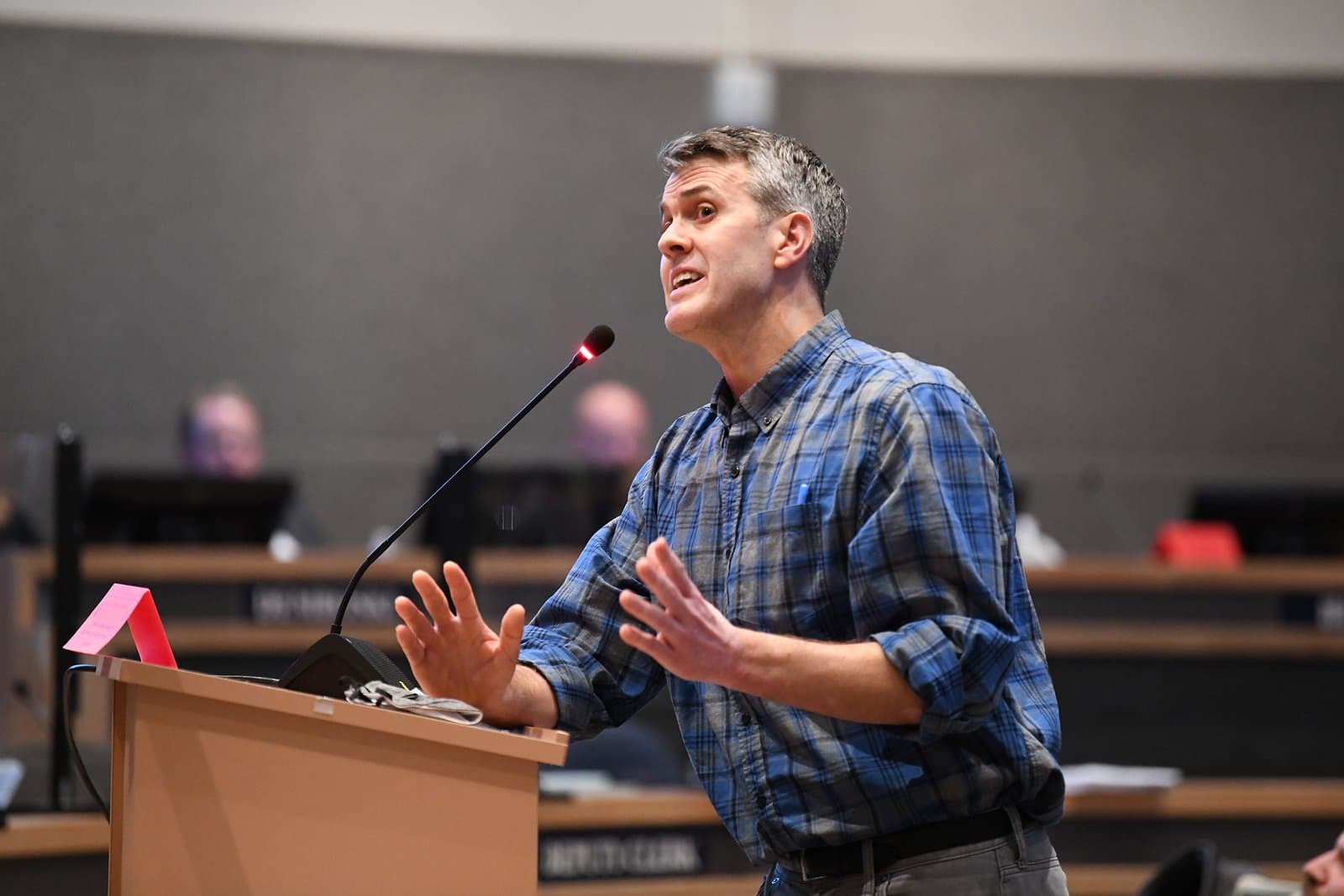 A man with short grey hair and a blue plaid shirt speaks passionately at a podium with a microphone, gesturing with both hands during an Anchorage Assembly meeting.