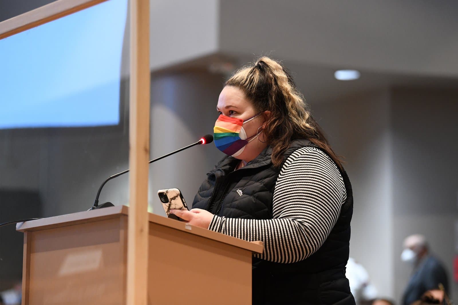 A woman wearing a rainbow-patterned face mask speaks into a microphone at an Anchorage Assembly meeting while holding a smartphone.