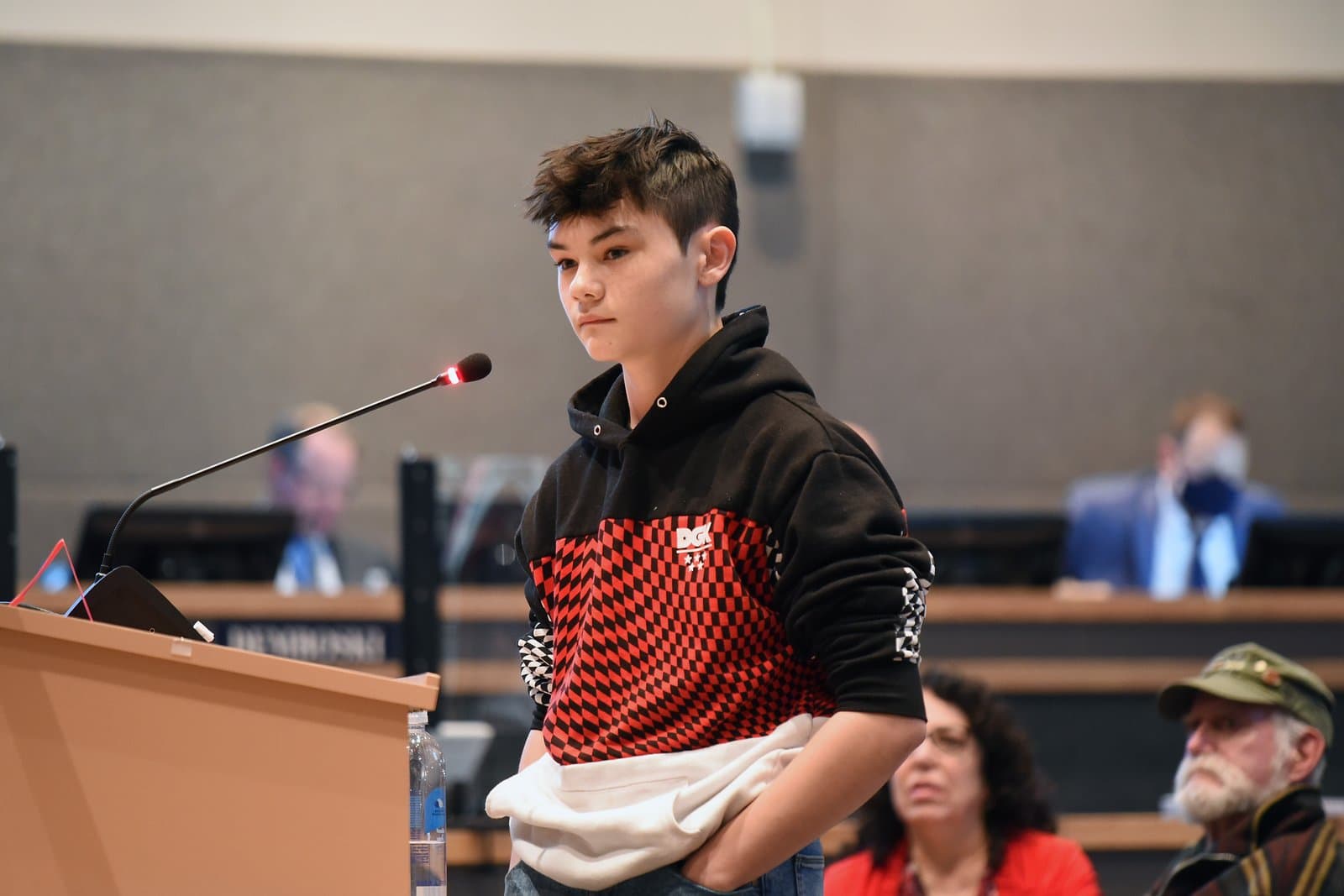 A young person with dark hair stands at a wooden podium, speaking into a microphone with a red light, during an Anchorage Assembly meeting. They are wearing a black, red, and white checkered hoodie, with other attendees visible in the blurred background.