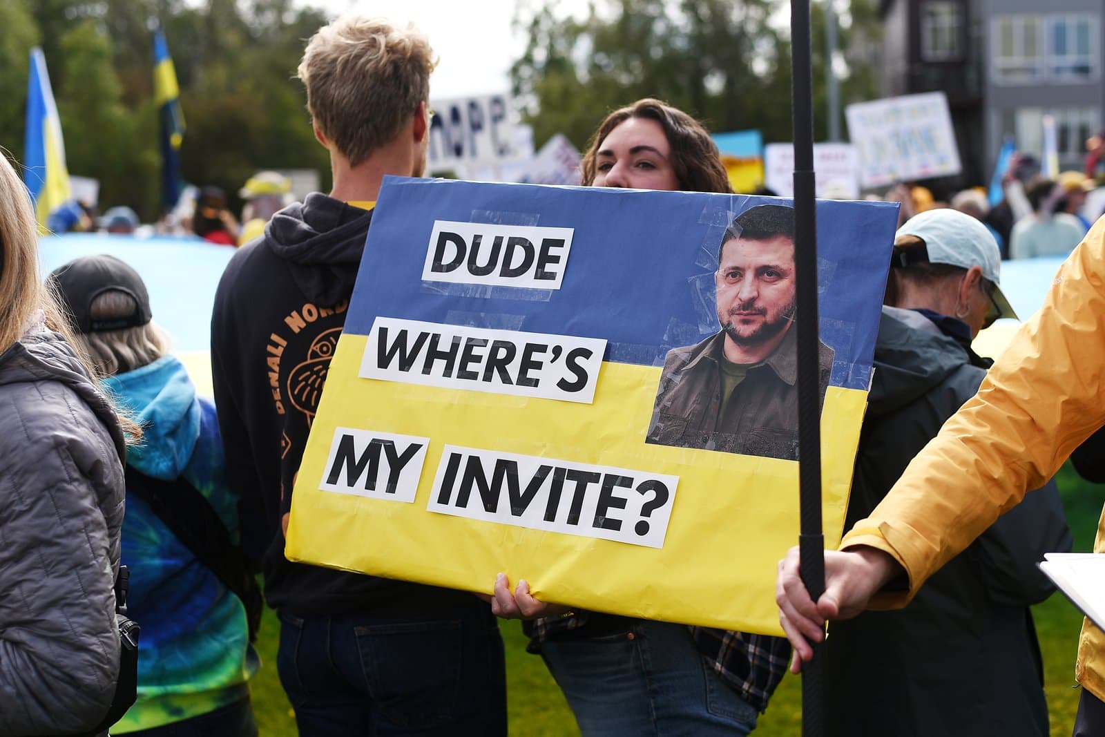 A protester holds a sign featuring Volodymyr Zelenskyy and the text Dude Where's My Invite during a pro-Ukraine demonstration in downtown Anchorage.