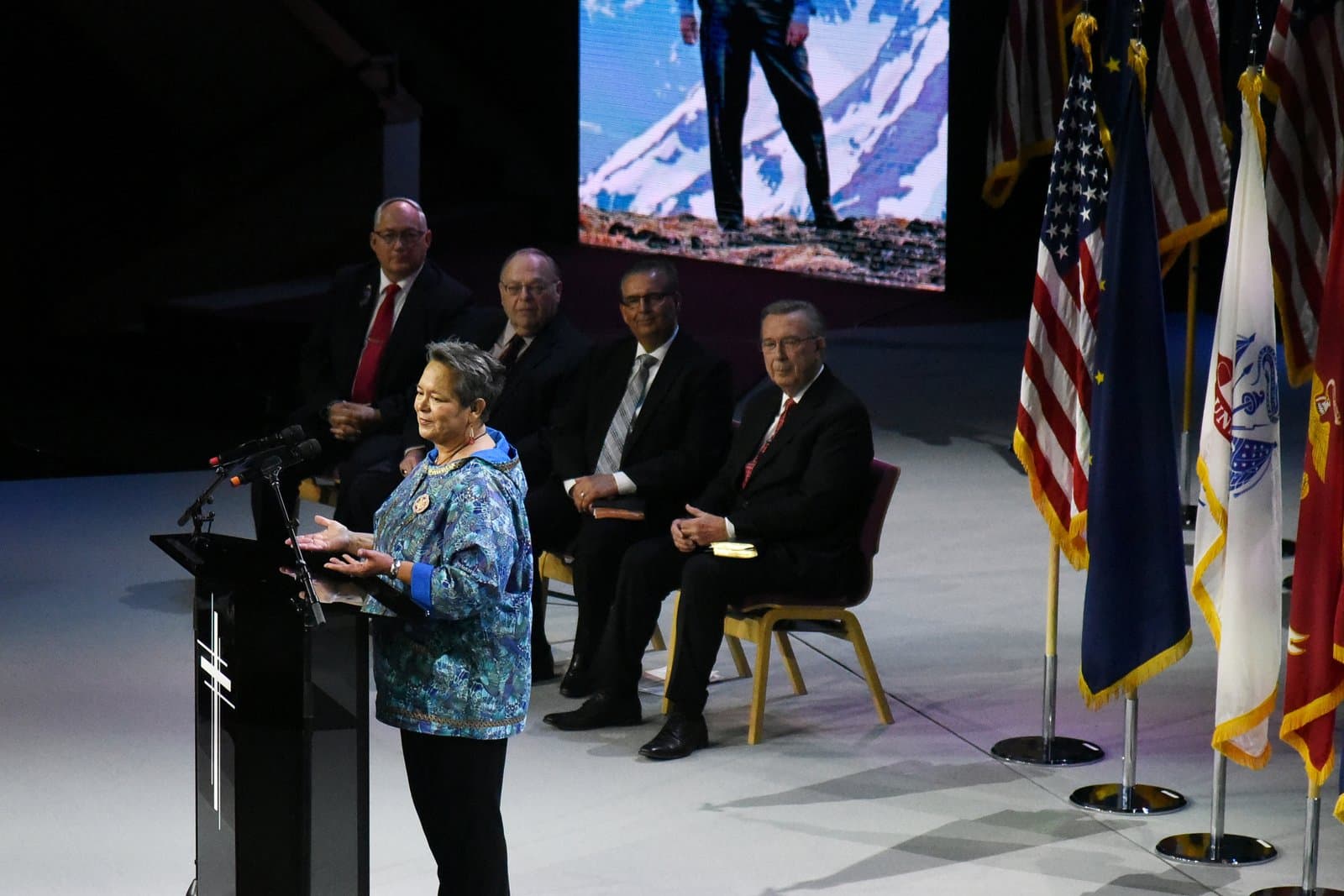 A woman speaks at a podium during the memorial service for Don Young, with seated officials and American flags in the background.