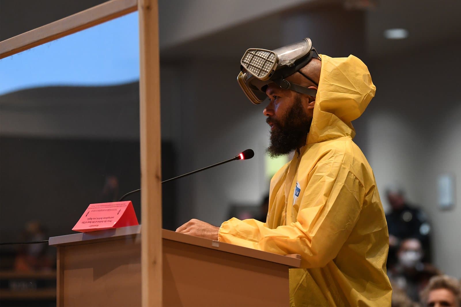 A man with a beard, wearing a yellow hazmat suit and a respirator mask, stands at a wooden podium speaking into a microphone at an indoor public meeting, with a clear barrier in front of him.
