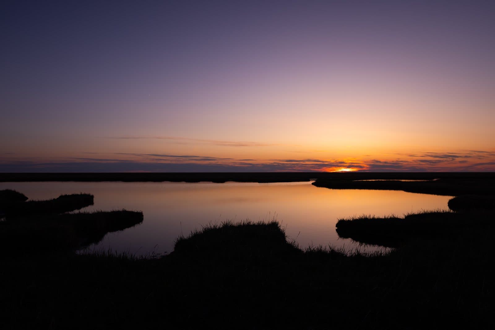 A vibrant sunset illuminates a calm tundra pond, casting a golden reflection across the water and silhouetting the surrounding grassy banks.
