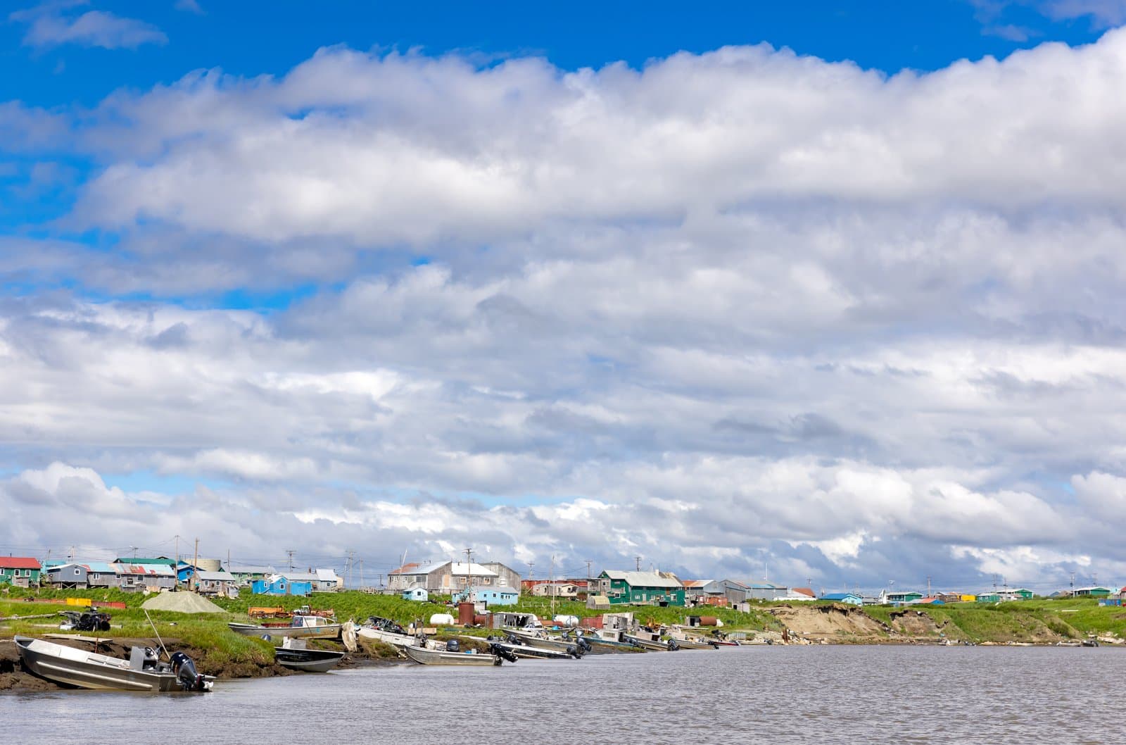 Small aluminum skiffs are moored along the muddy riverbank in the village of Chevak under a bright, cloud-filled summer sky.